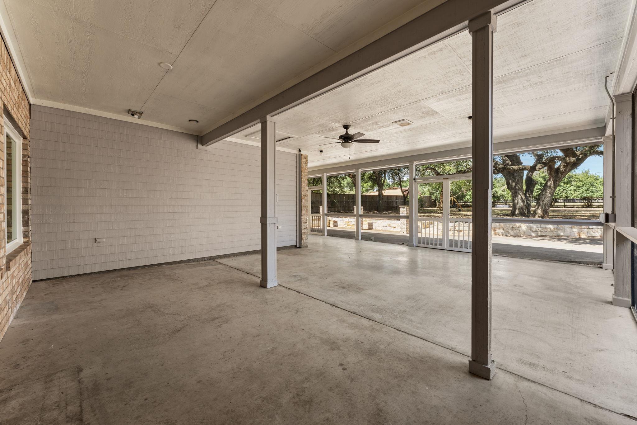 Covered concrete patio with a ceiling fan, white railings, and mature trees beyond.