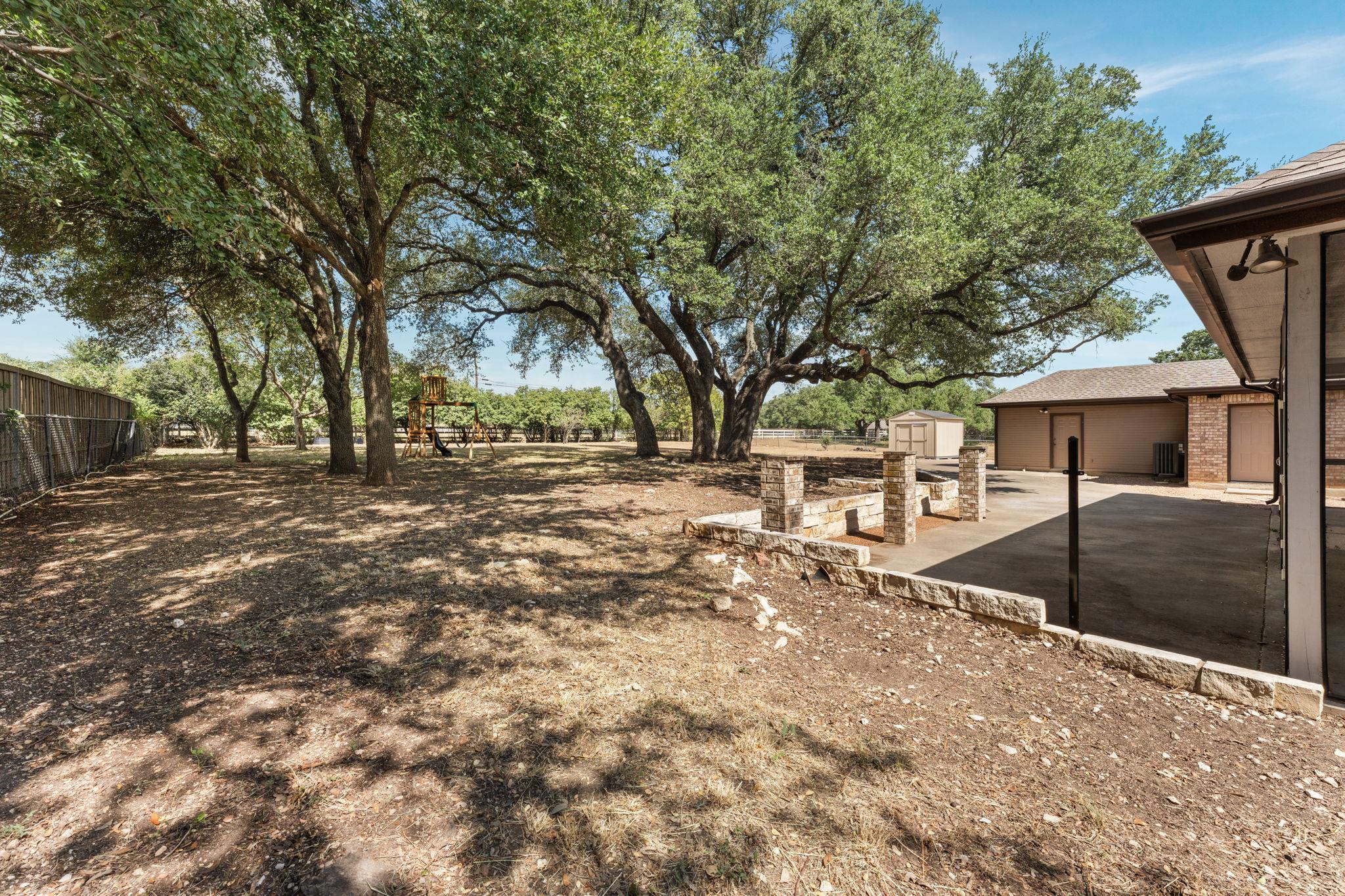 Shaded backyard with mature oak trees, a playset, and a covered patio.