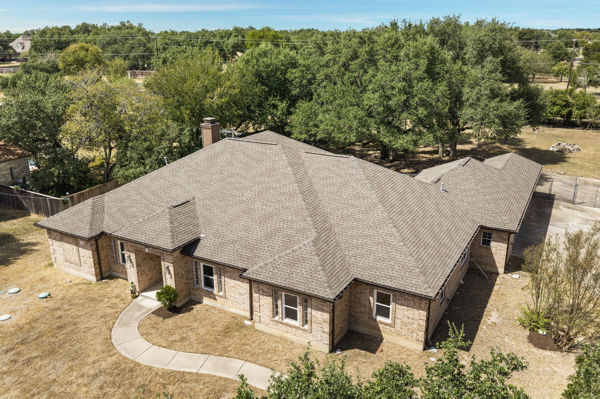 Aerial view of a brick home with a broad shingle roof, dry lawn, and mature trees.