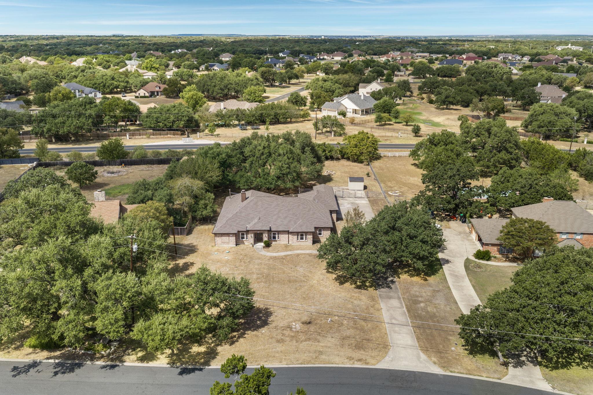 Aerial view of a brick home on a large tree-shaded lot, with a road and neighborhood beyond.