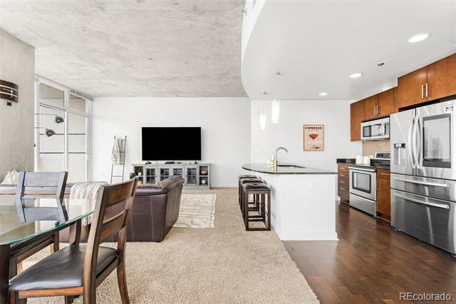 Open-plan condo living room and kitchen with a white island, stainless steel appliances, and wood cabinets.