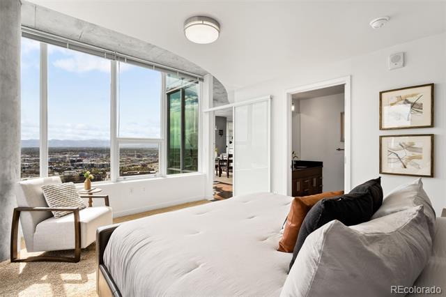 Bedroom with floor-to-ceiling windows overlooking Denver, a large bed, and a sitting chair.
