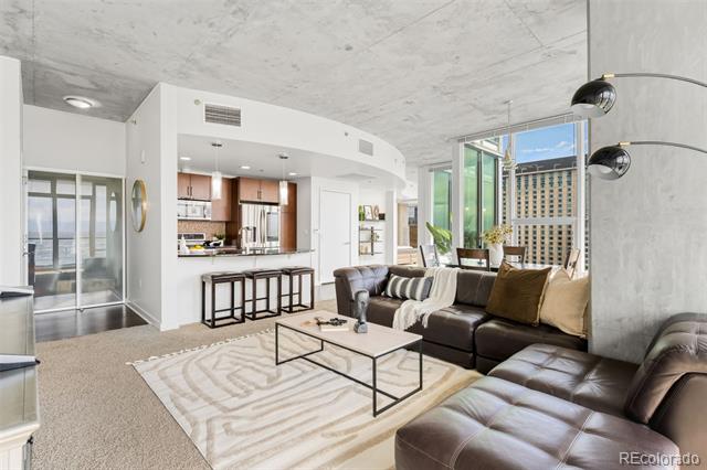 Open-plan condo living room with a leather sectional, exposed concrete ceiling, and floor-to-ceiling city-view windows.