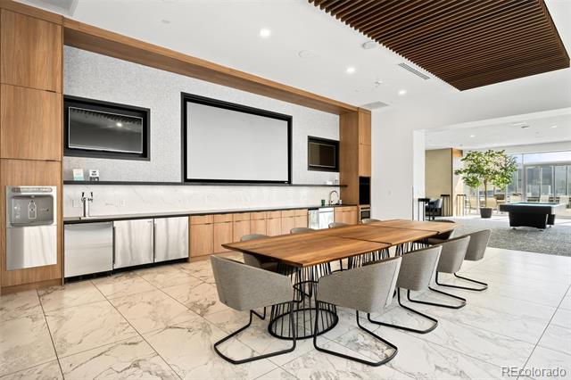 Furnished shared dining area with a long wood table beside a kitchenette and bright lounge.