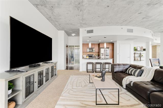Condo living room with a wall-mounted television, dark sectional, glass coffee table, and open kitchen.