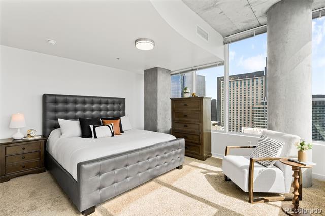 Bedroom with a gray upholstered bed, tufted black headboard, and floor-to-ceiling windows overlooking city buildings.