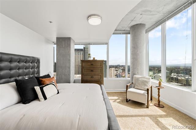 High-rise bedroom with a tufted headboard and armchair beside floor-to-ceiling windows overlooking the city.