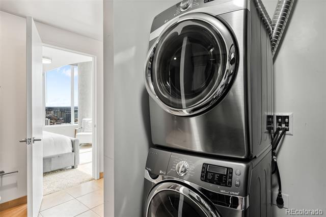 Stacked stainless-steel washer and dryer beside a doorway to a bright bedroom.