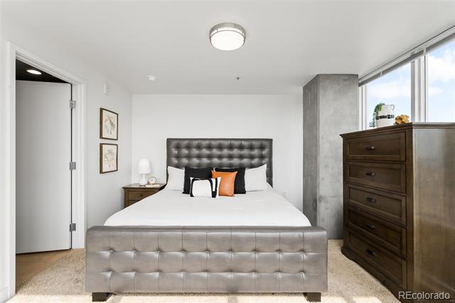 Bedroom with a tufted gray bed, orange accent pillow, dark wood dresser, and tall windows.