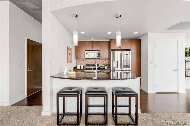 Condominium kitchen with a dark stone island, three bar stools, and stainless steel appliances.