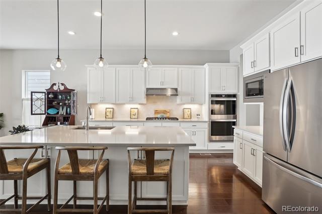 White kitchen with a large island, three wooden stools, stainless steel appliances, and dark hardwood floors.