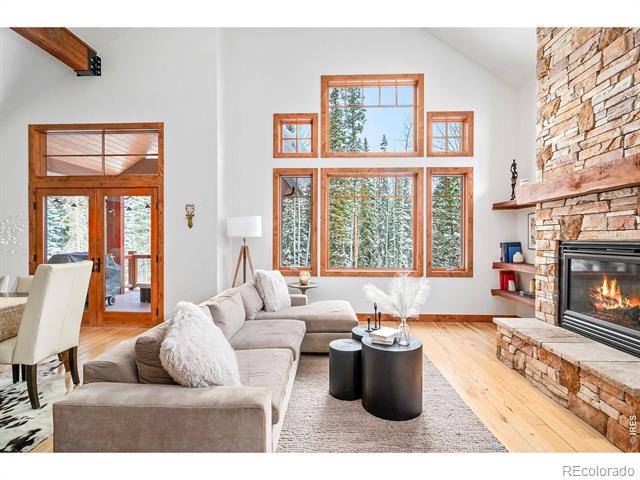Living room with a gray sectional, tall forest-view windows, and a stone fireplace.