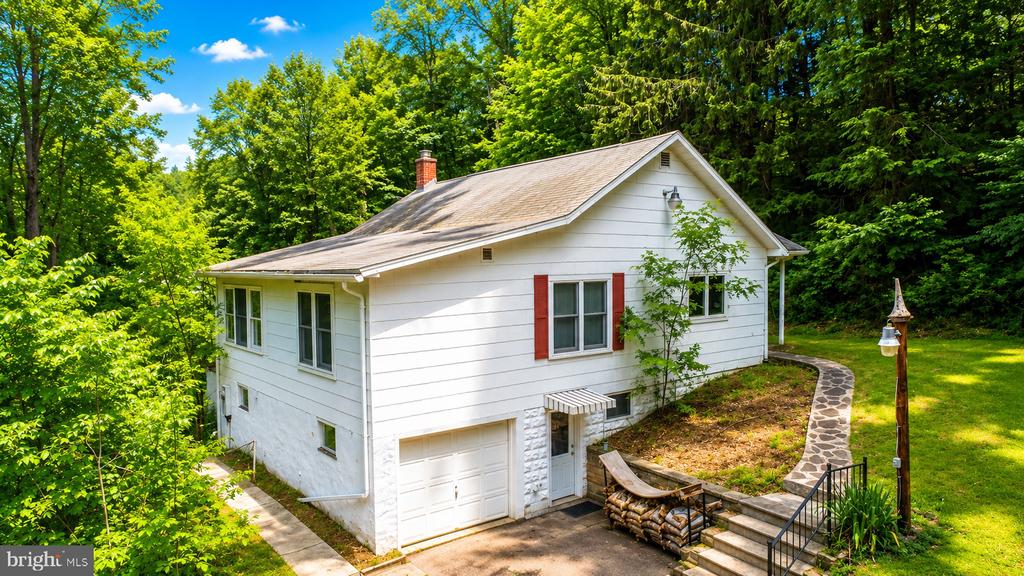 White split-level house with red shutters, an attached garage, and a curved stone path in a wooded yard.