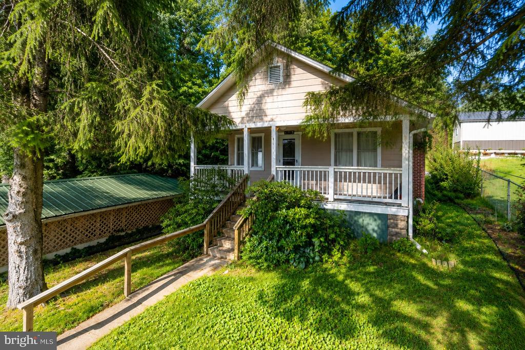 Light-colored house with a covered porch, wooden ramp, and mature evergreen trees.