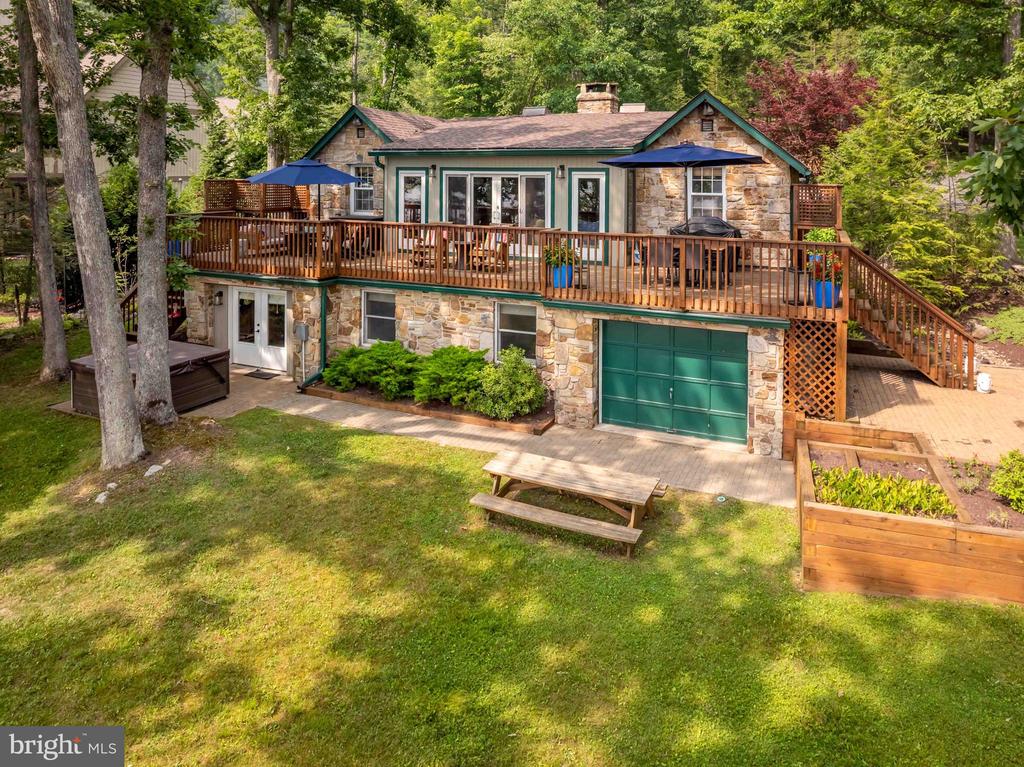 Stone-and-siding home with a raised wooden deck, patio umbrellas, and green garage door among trees.