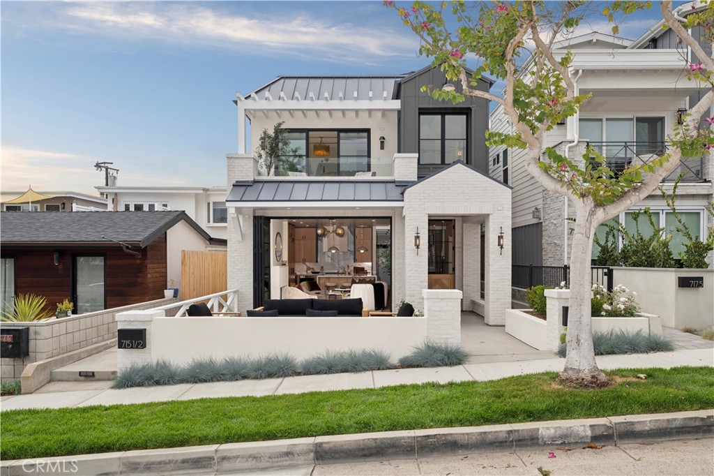 Modern white-brick home with black-framed windows, covered patio seating, and a flowering tree by the walkway.