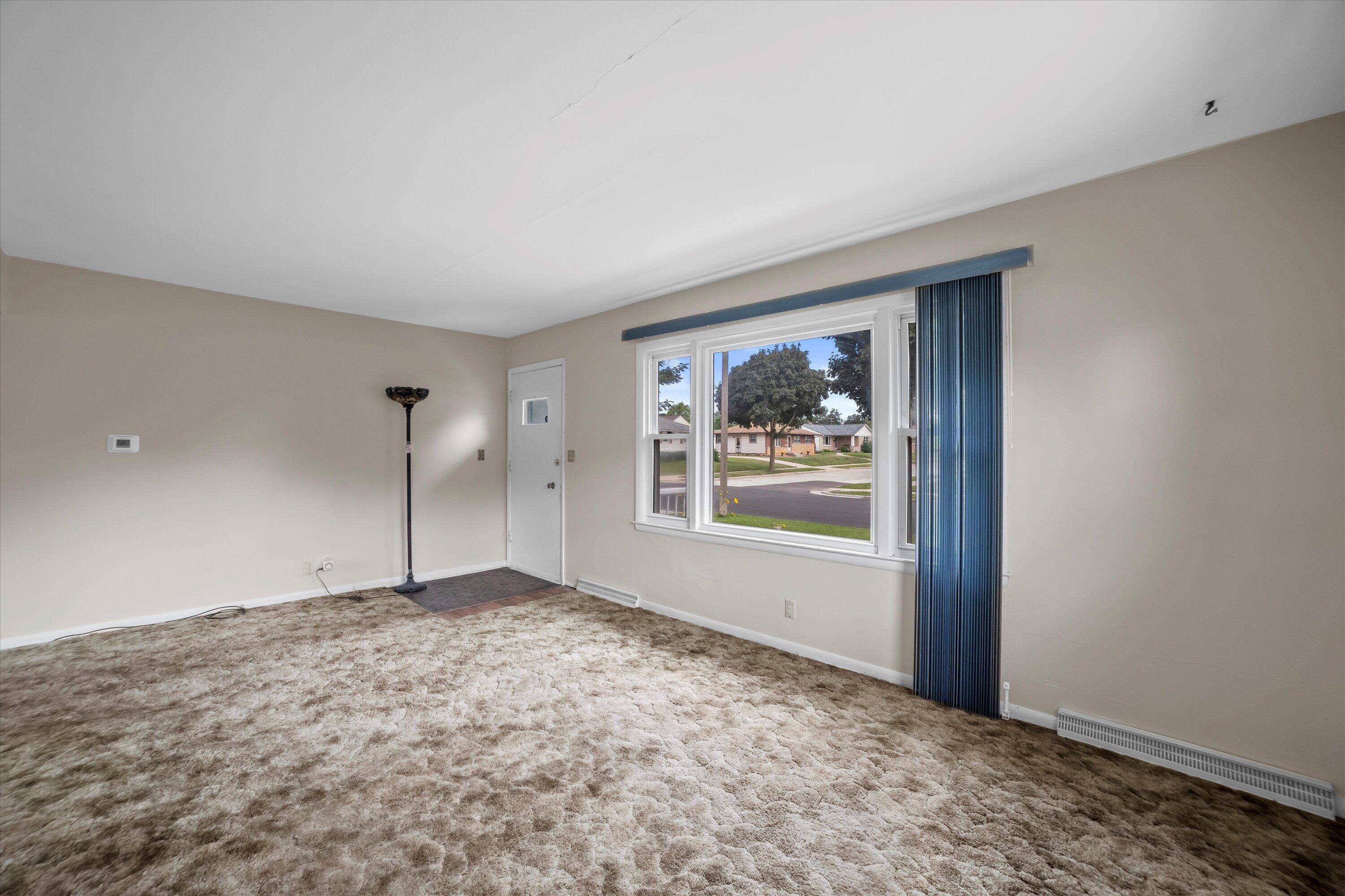 Empty living room with plush beige carpet and a large window overlooking a residential street.