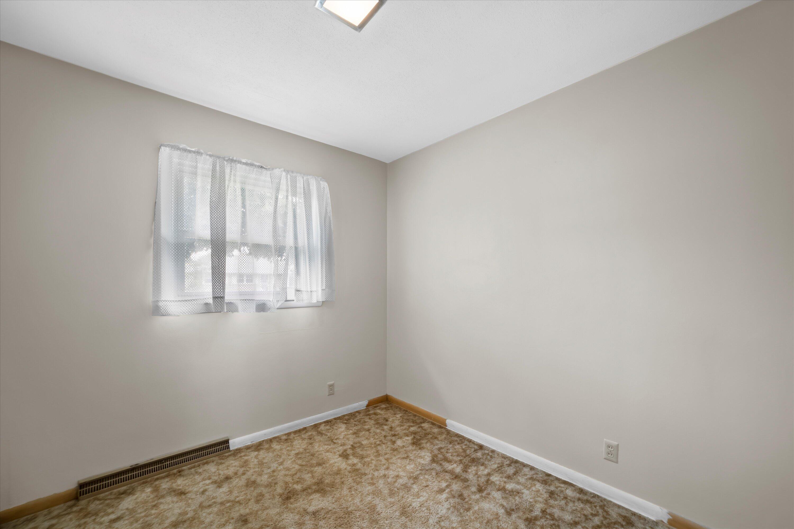 Empty bedroom with tan carpet, a curtained window, and a floor-level heating vent.