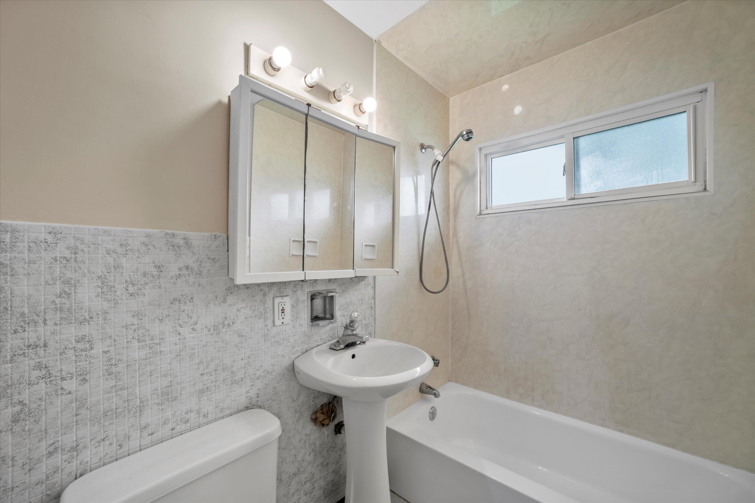 Bathroom with a white tub, pedestal sink, mirrored cabinet, and high frosted-glass window.