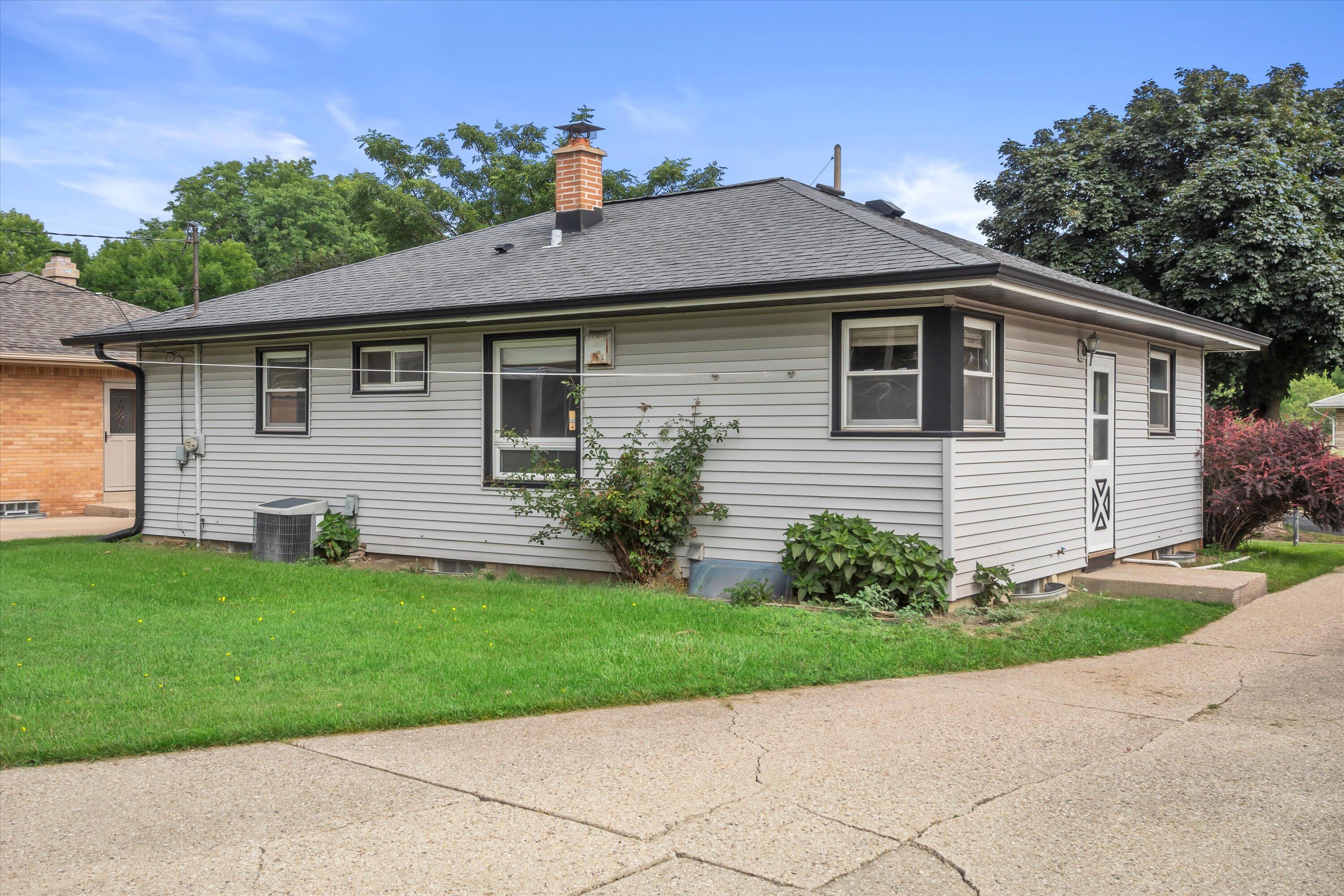 Single-story gray-sided house with a bay window, brick chimney, and green lawn.