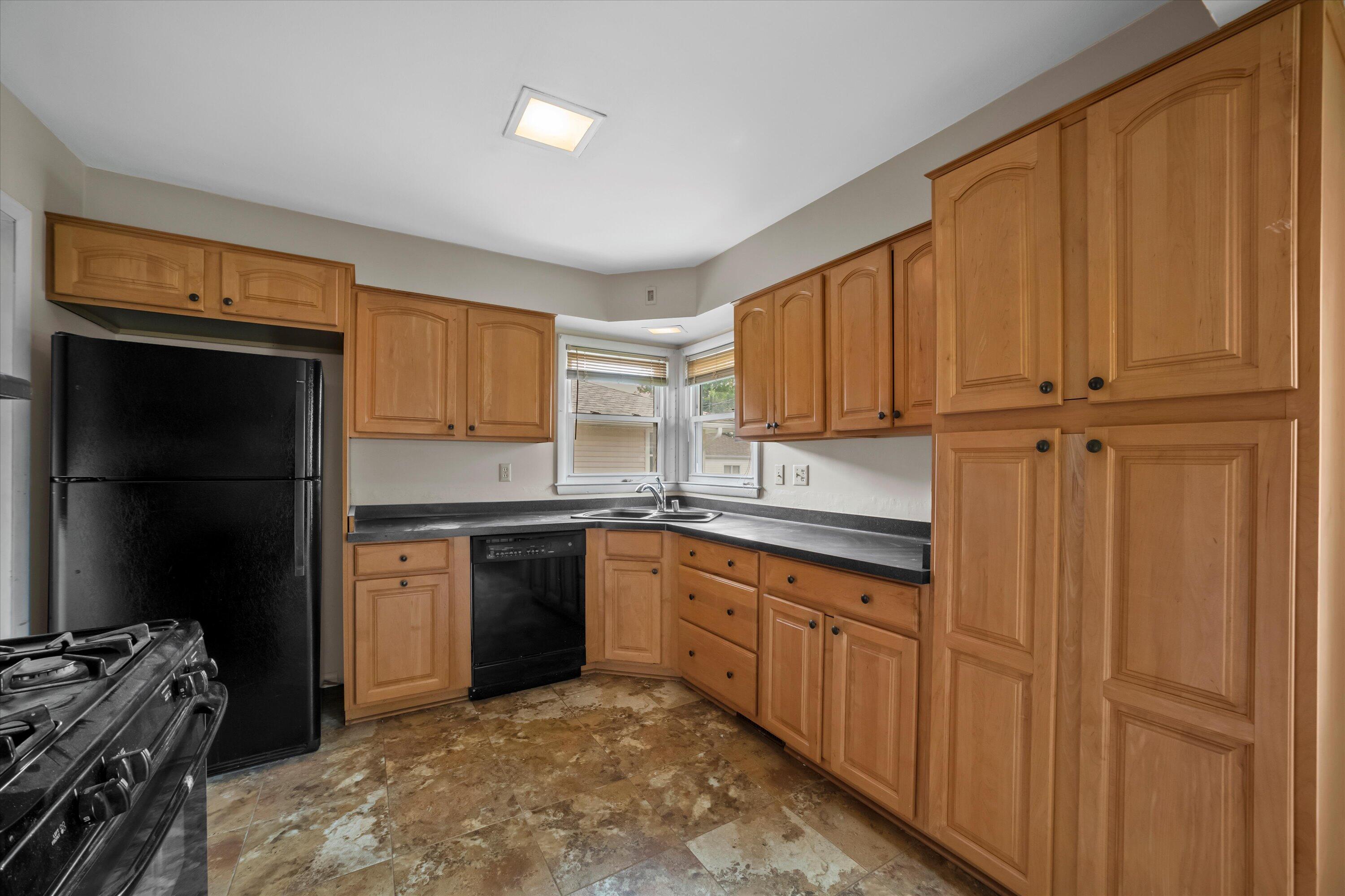 Kitchen with honey-colored cabinets, dark countertops, black appliances, and a corner sink beneath two windows.