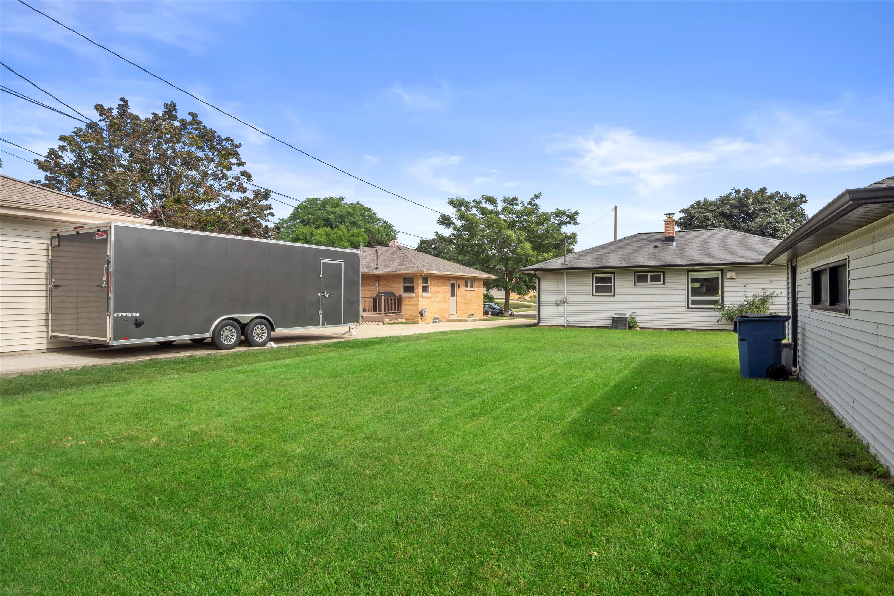 Green backyard between homes, with a large gray enclosed trailer parked beside the lawn.