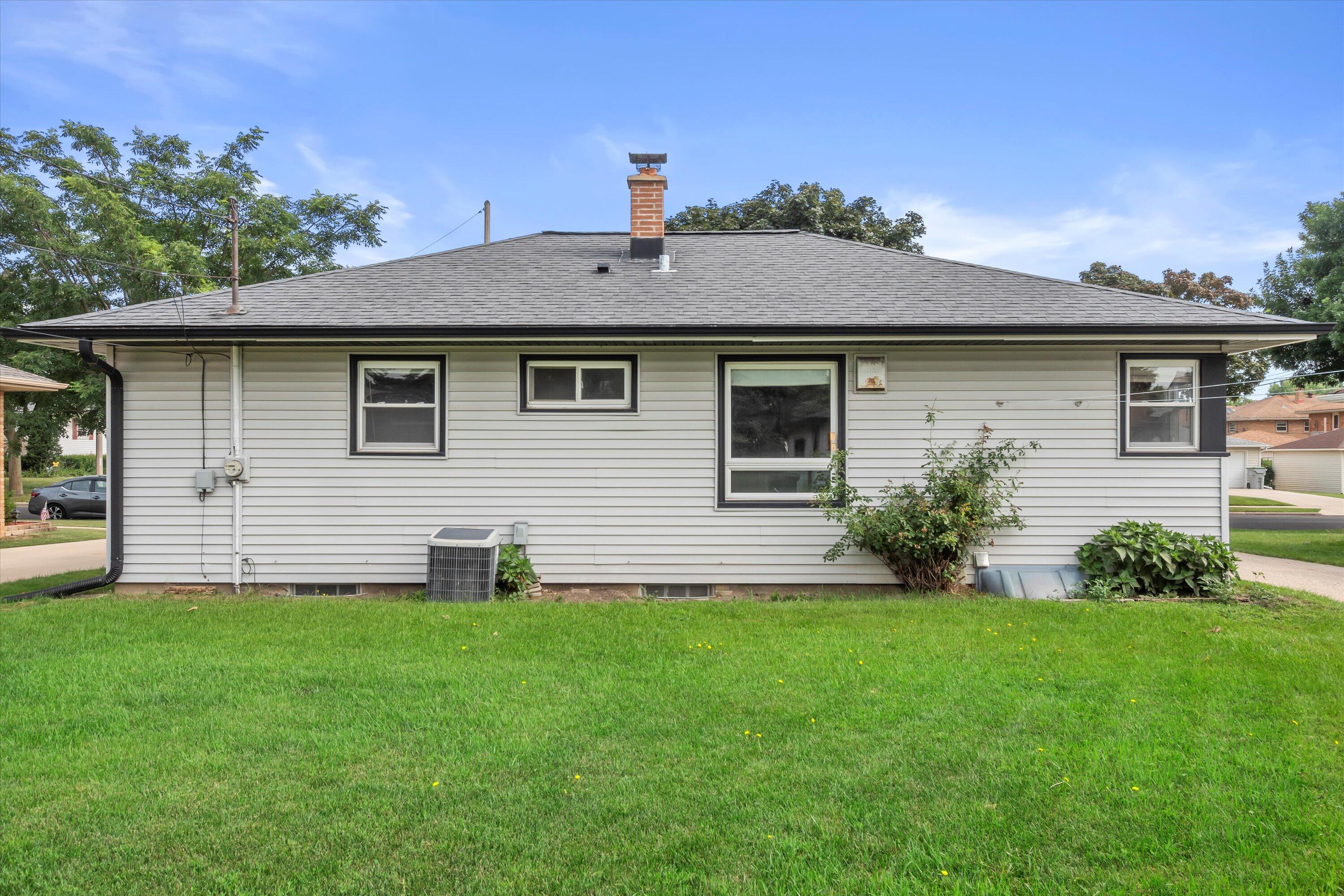 Side of a gray-sided single-story house with a brick chimney, shrubs, and a green lawn.