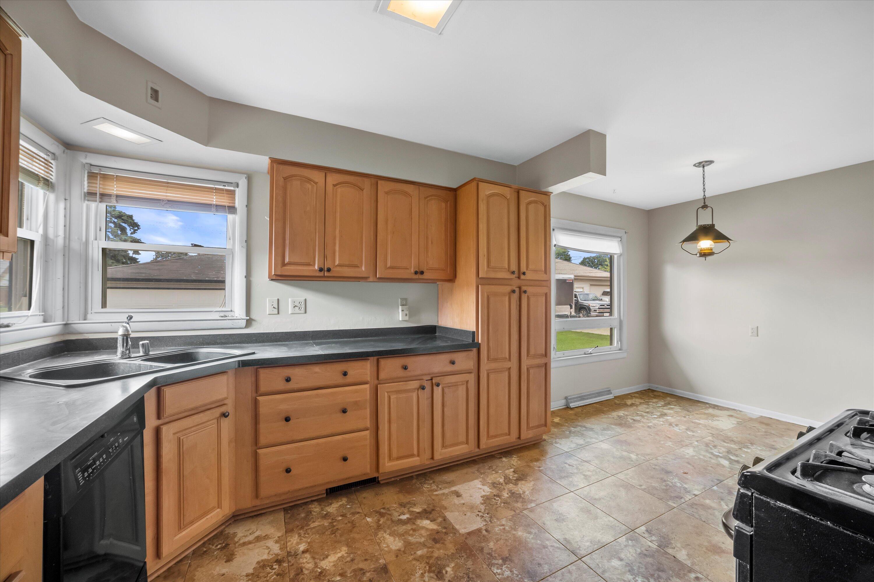 Kitchen with honey-colored cabinets, dark countertops, tiled floor, and windows above the sink.