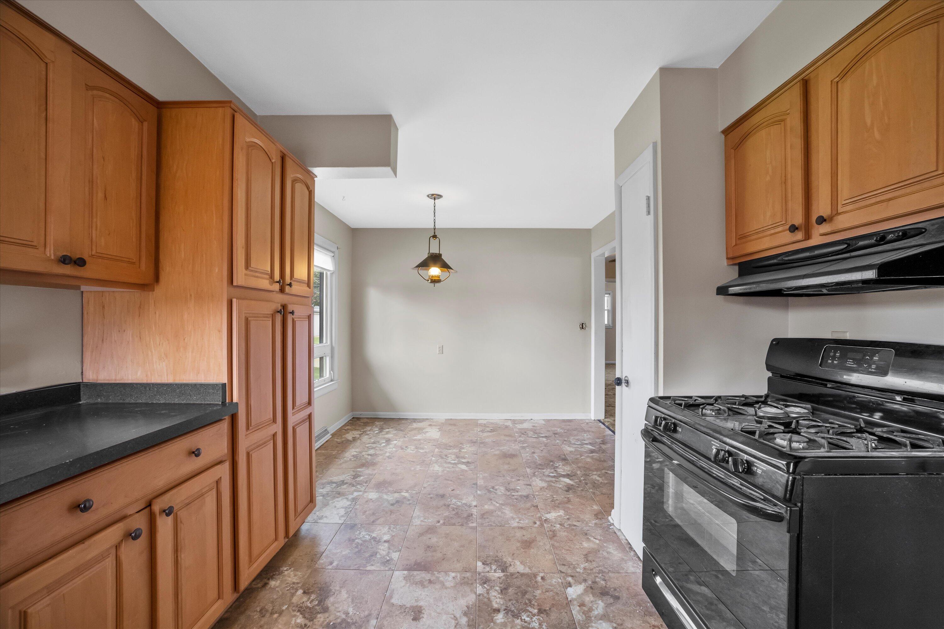 Empty kitchen with wood cabinets, black gas range, and tile floor opening to a dining area.