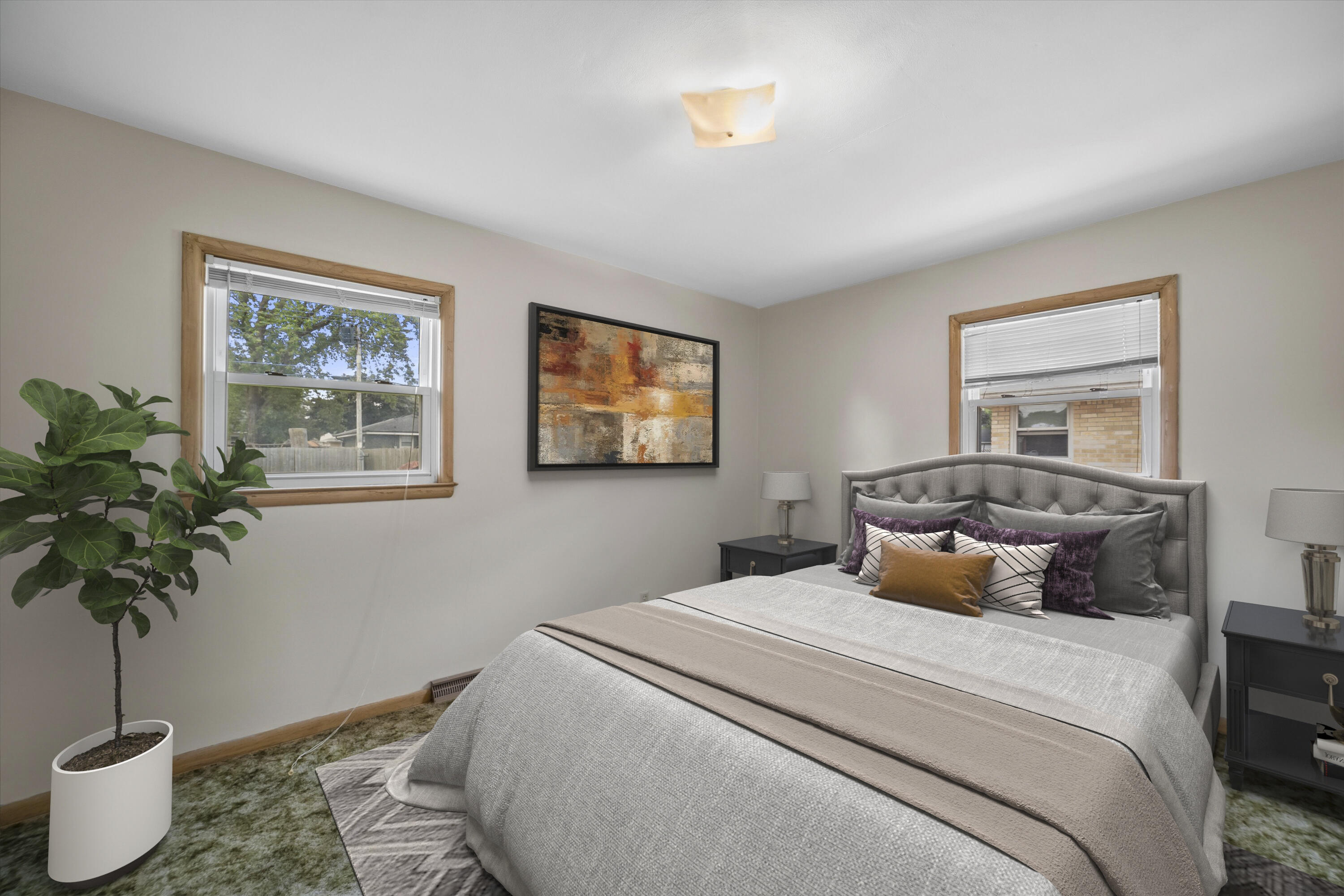 Staged bedroom with a gray upholstered bed, colorful pillows, wood-trimmed windows, and a potted tree.