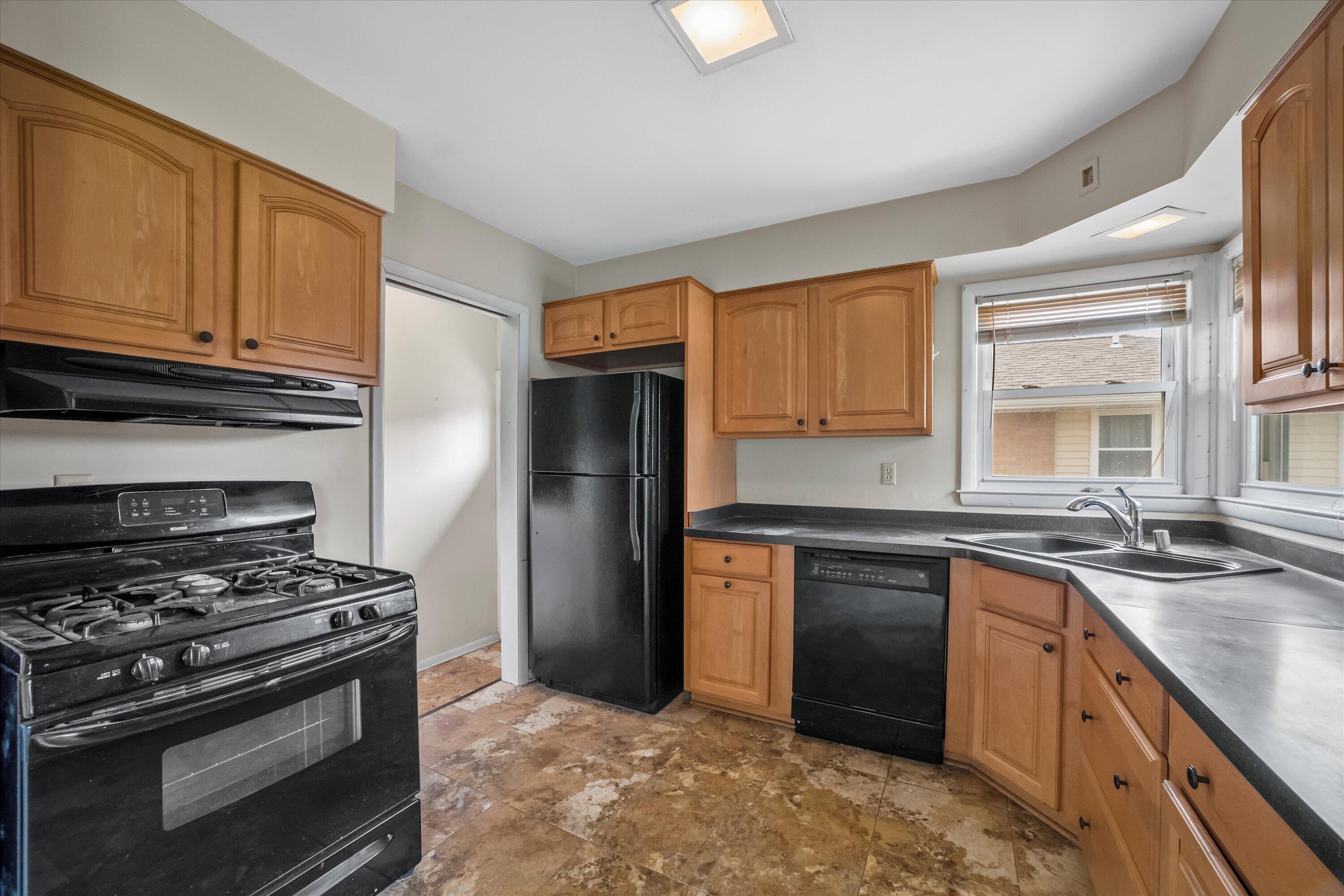 Kitchen with wood cabinets, dark counters, black appliances, and a double sink beneath windows.