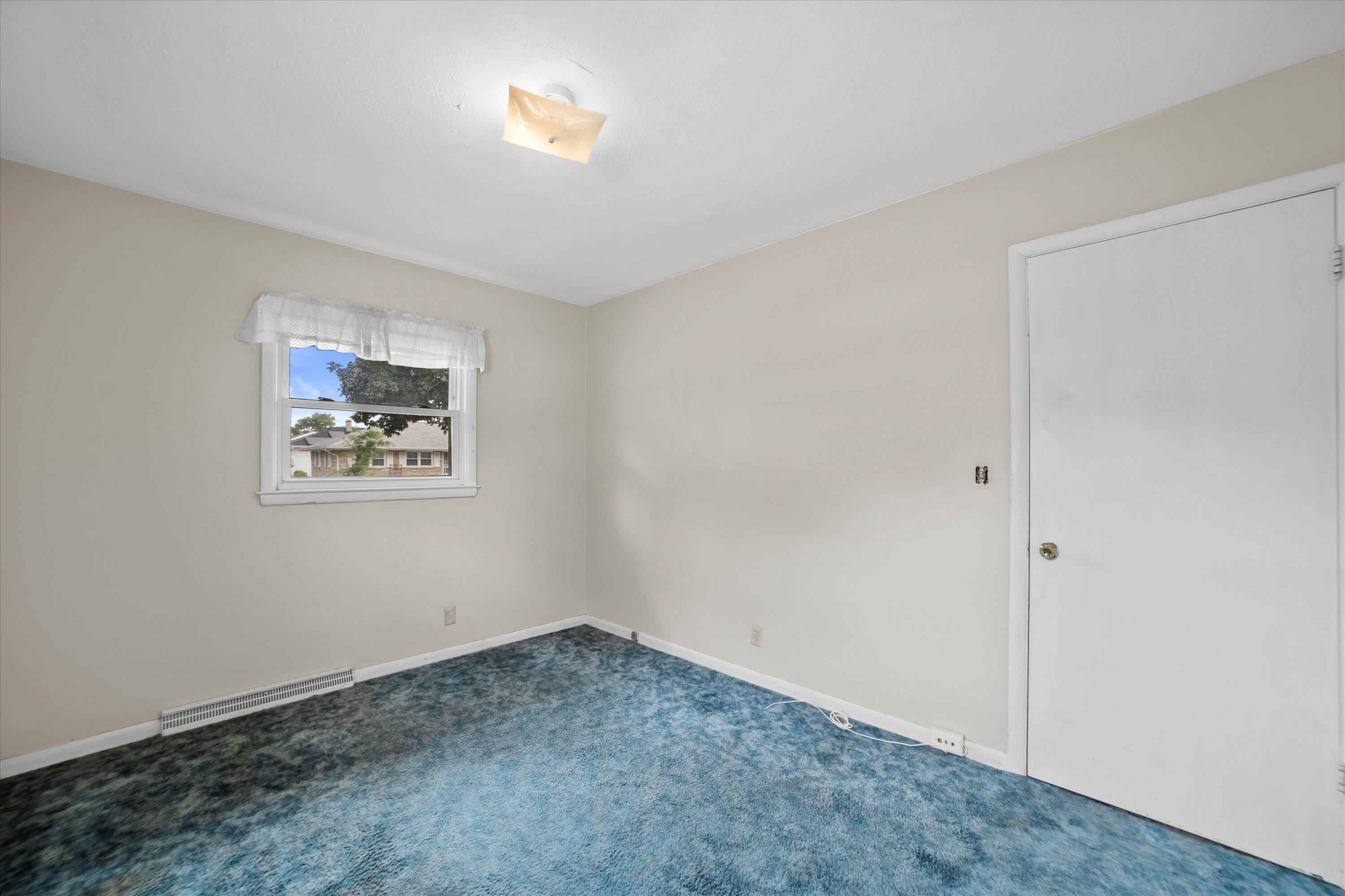 Empty bedroom with blue patterned carpet, beige walls, and a window with a white valance.