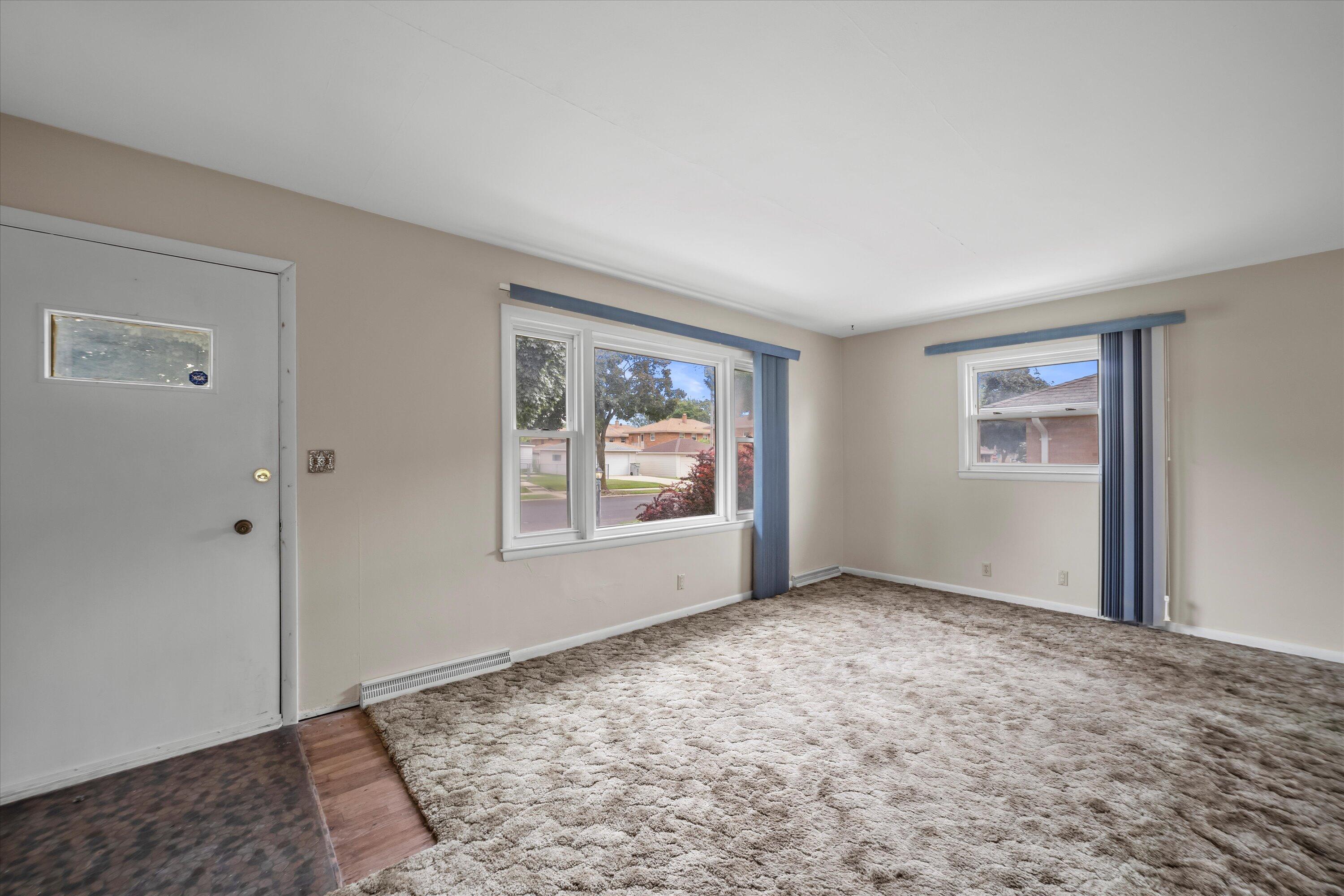 Empty living room with a gray entry door, large carpet, and windows dressed with blue vertical blinds.