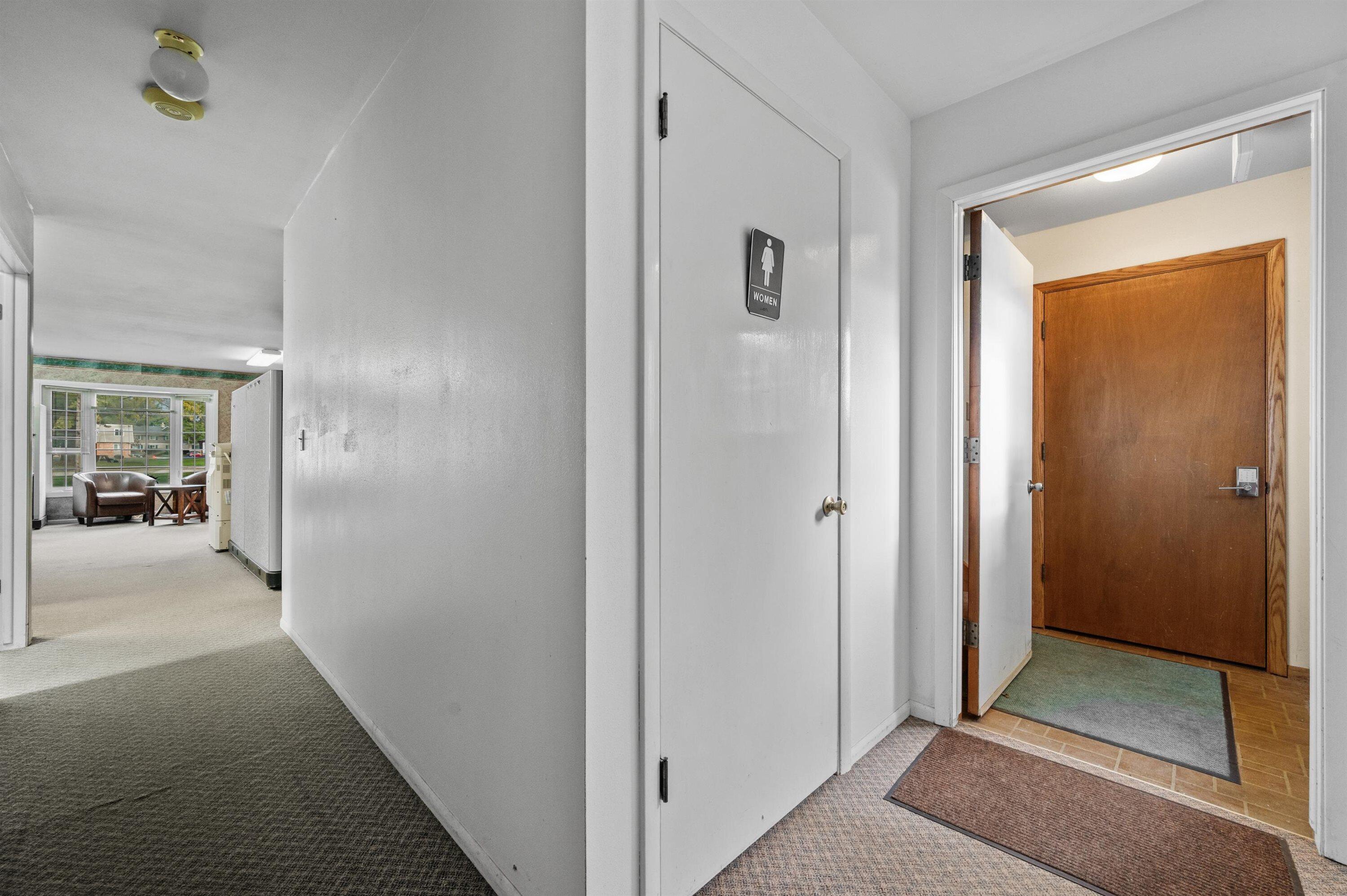 Hallway with a women’s restroom door, wooden entrance door, and lounge seating by a window.