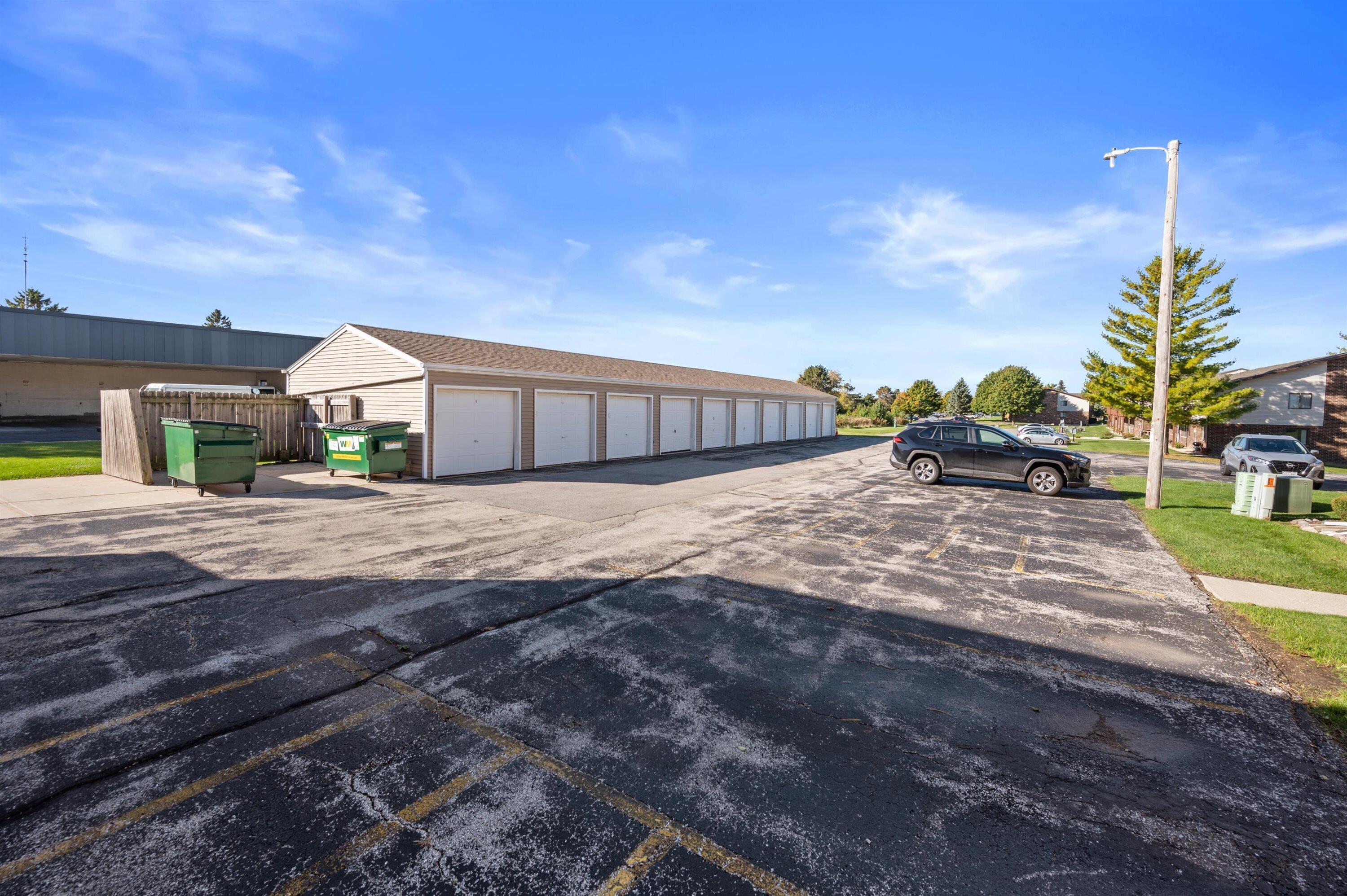 Row of beige garage units beside a cracked parking lot, with green dumpsters and parked cars.