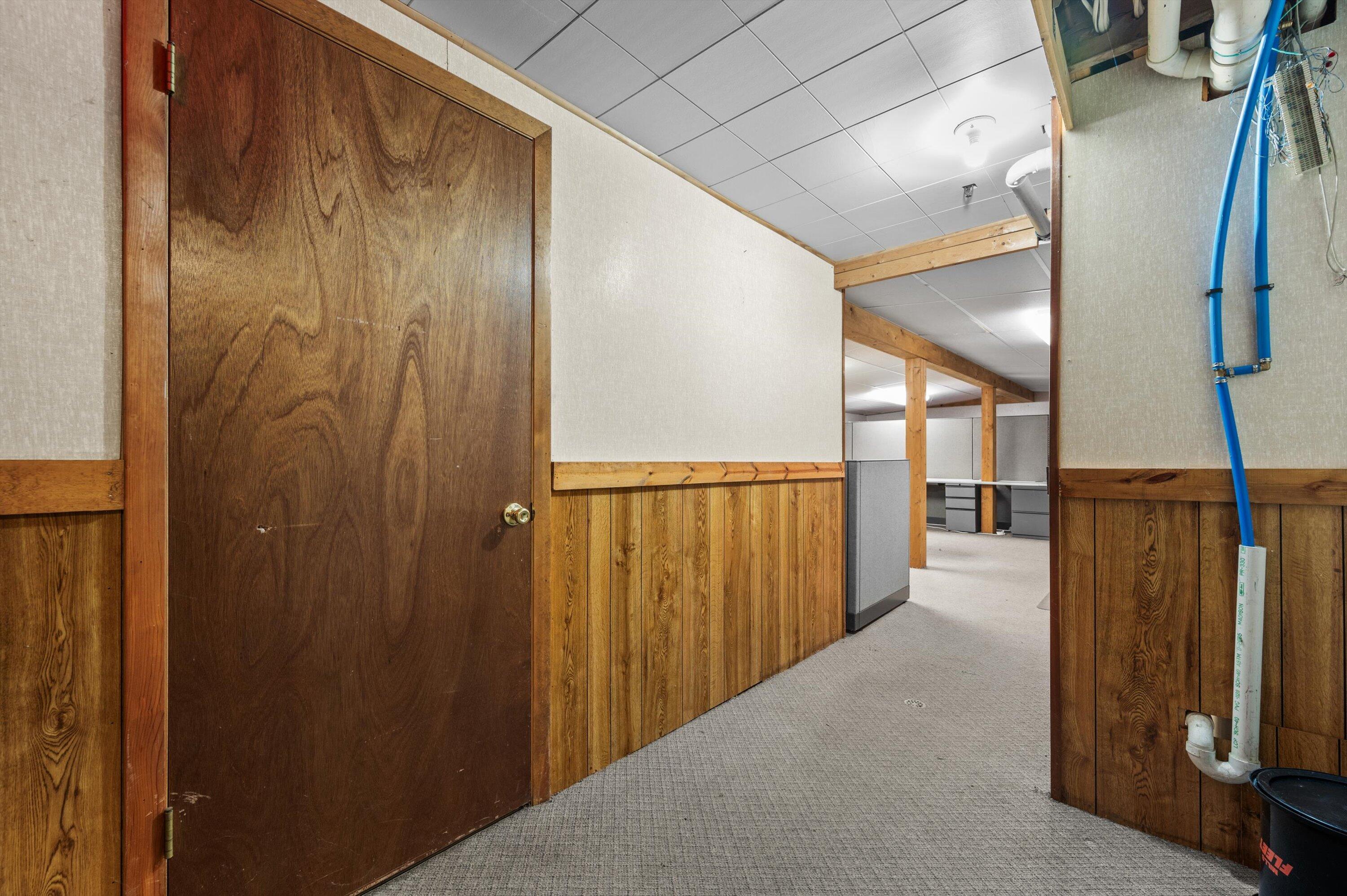 Vacant office hallway with wood-paneled walls, gray carpet, and cubicles beyond an open doorway.