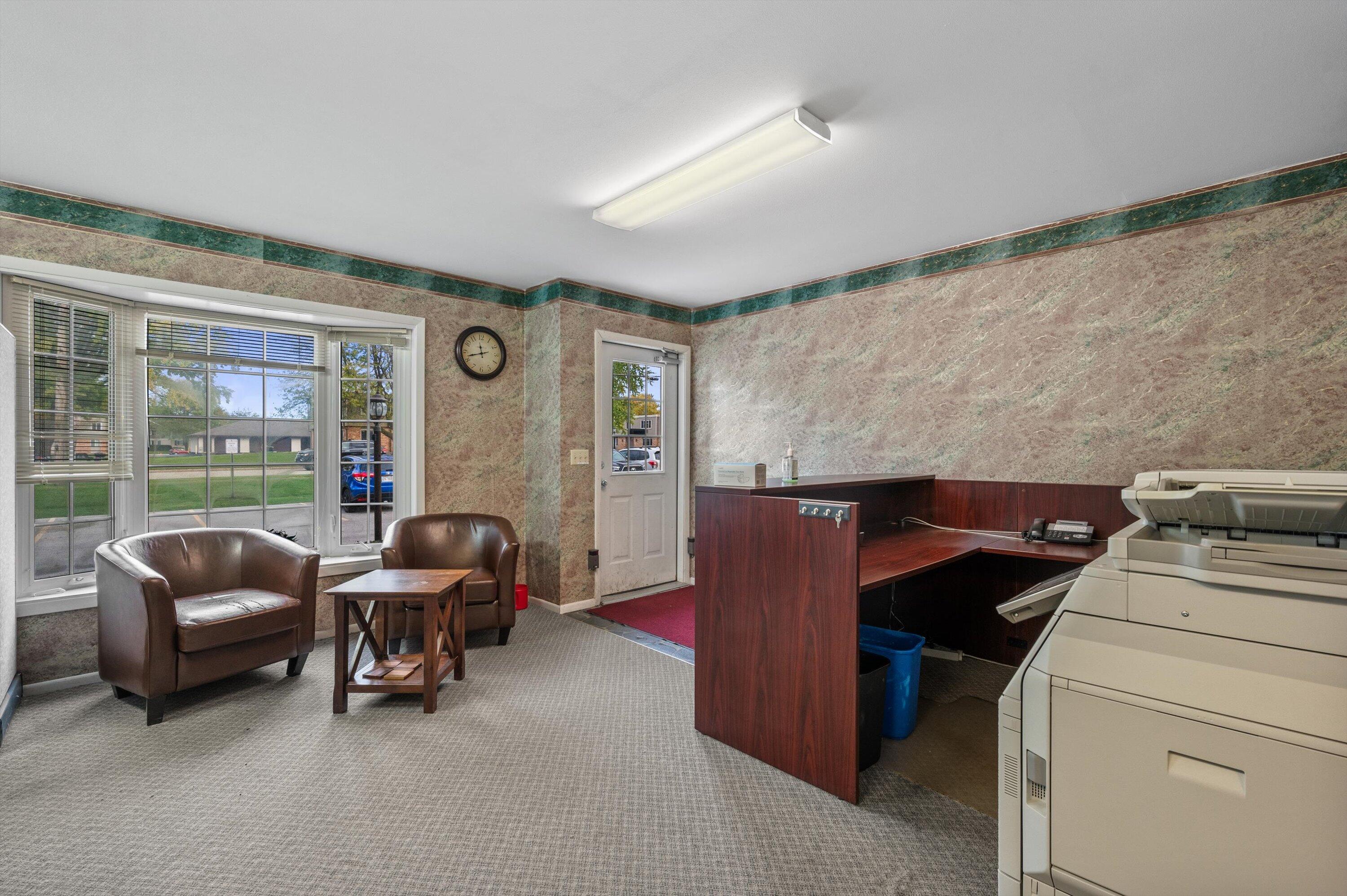 Office reception area with a dark wood counter, copier, brown armchairs, and large windows.