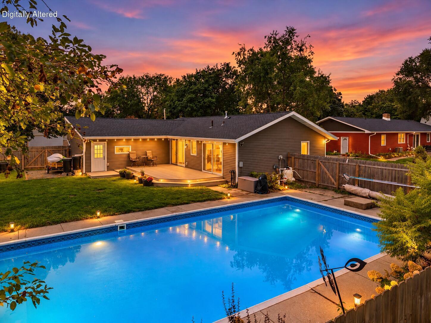 Backyard swimming pool beside a single-story home, deck, and fenced patio at dusk.