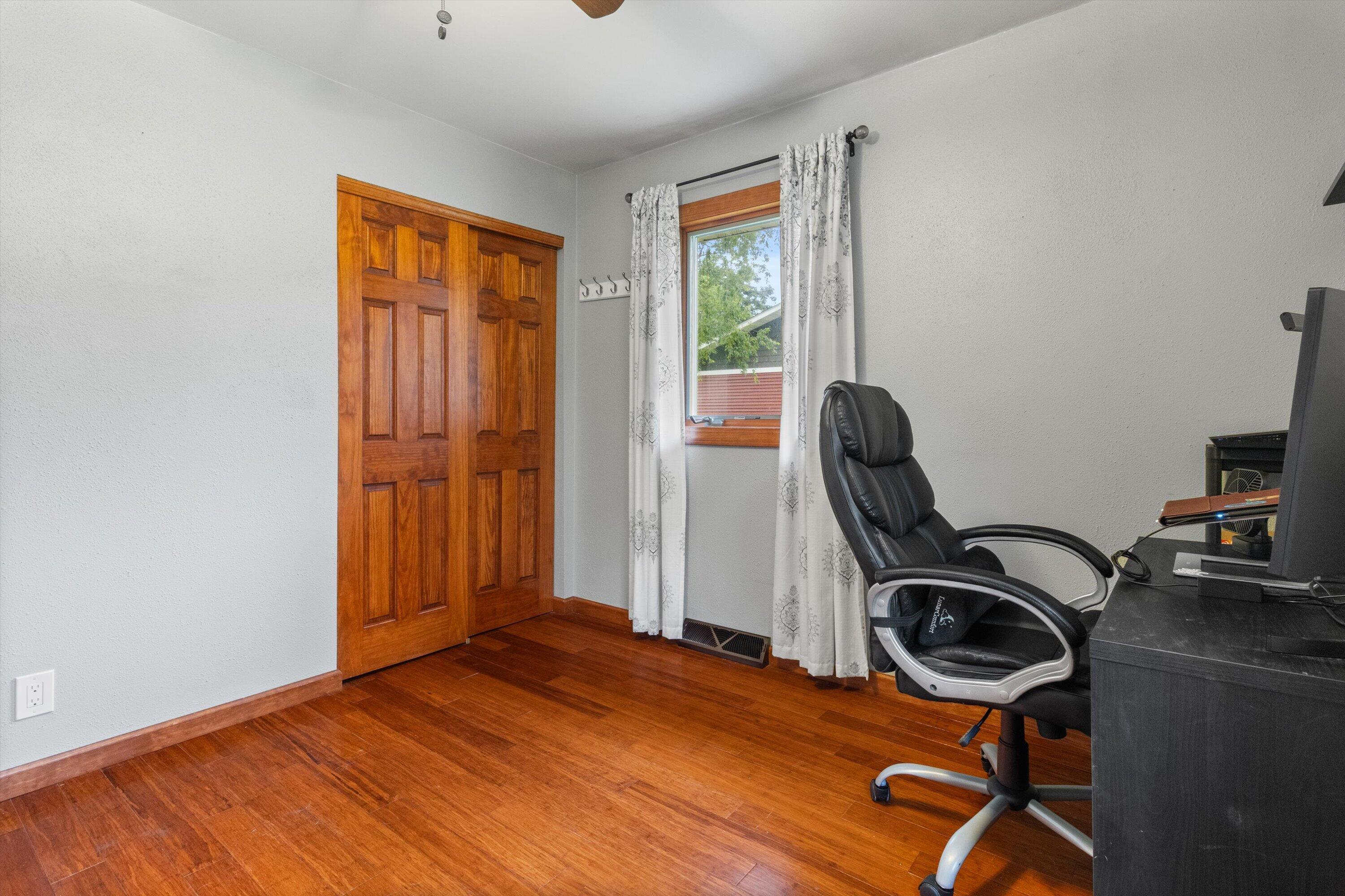 Small office with reddish hardwood floors, wooden closet doors, and a black desk chair by a curtained window.