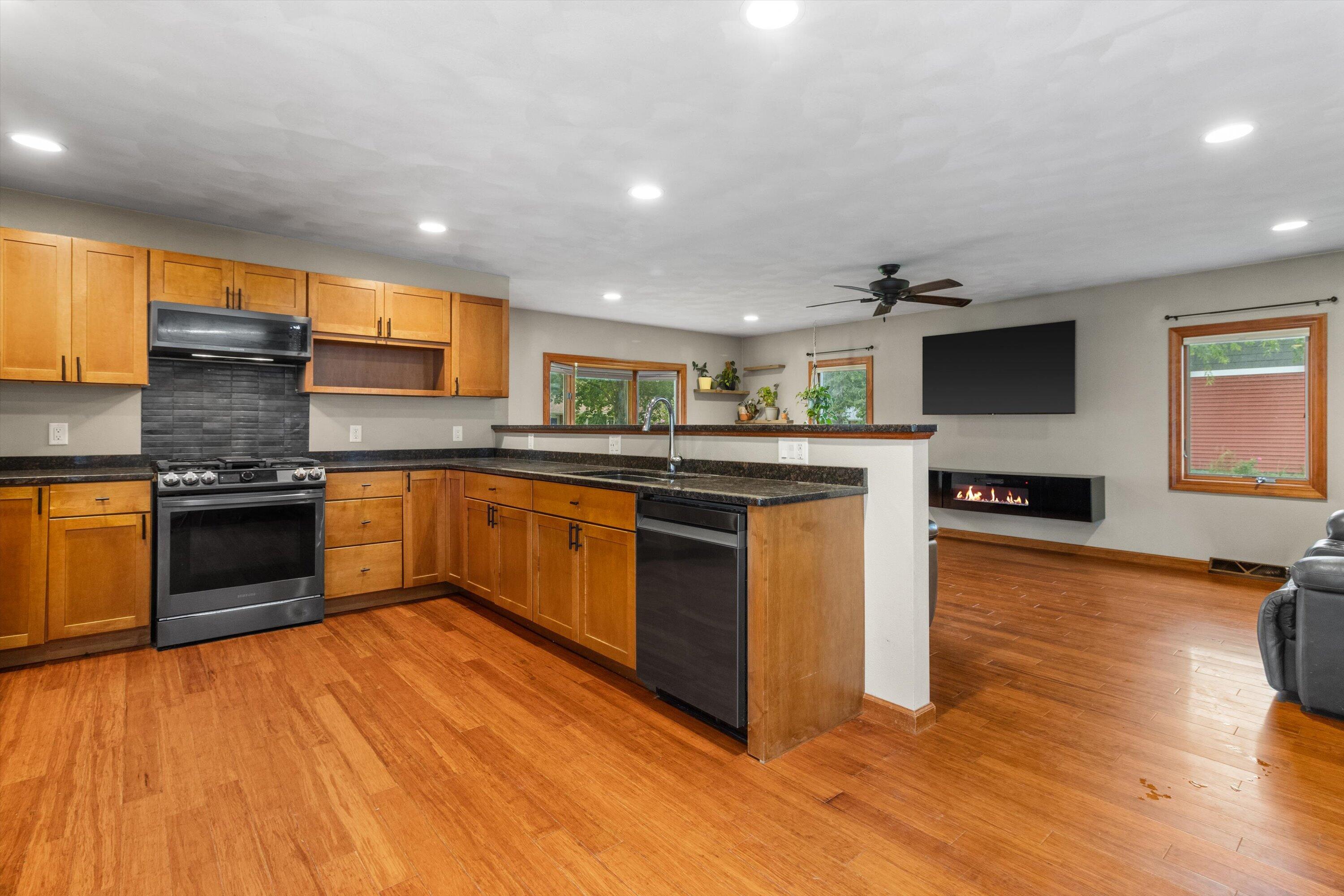 Open kitchen with warm wood cabinets, dark countertops, and a peninsula adjoining a living area with a fireplace.