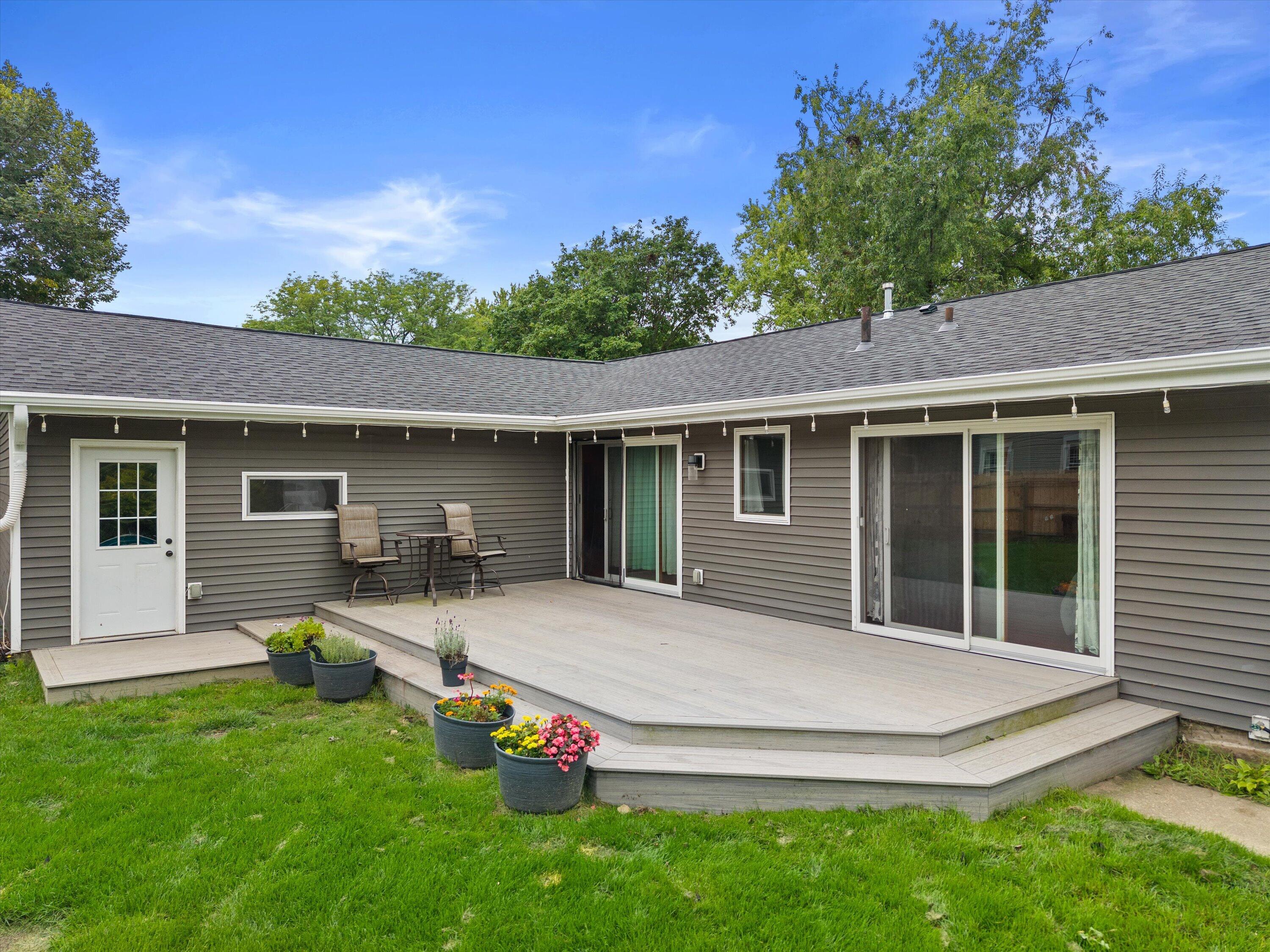 Gray-sided home with a raised deck, sliding glass doors, patio seating, and colorful potted flowers.