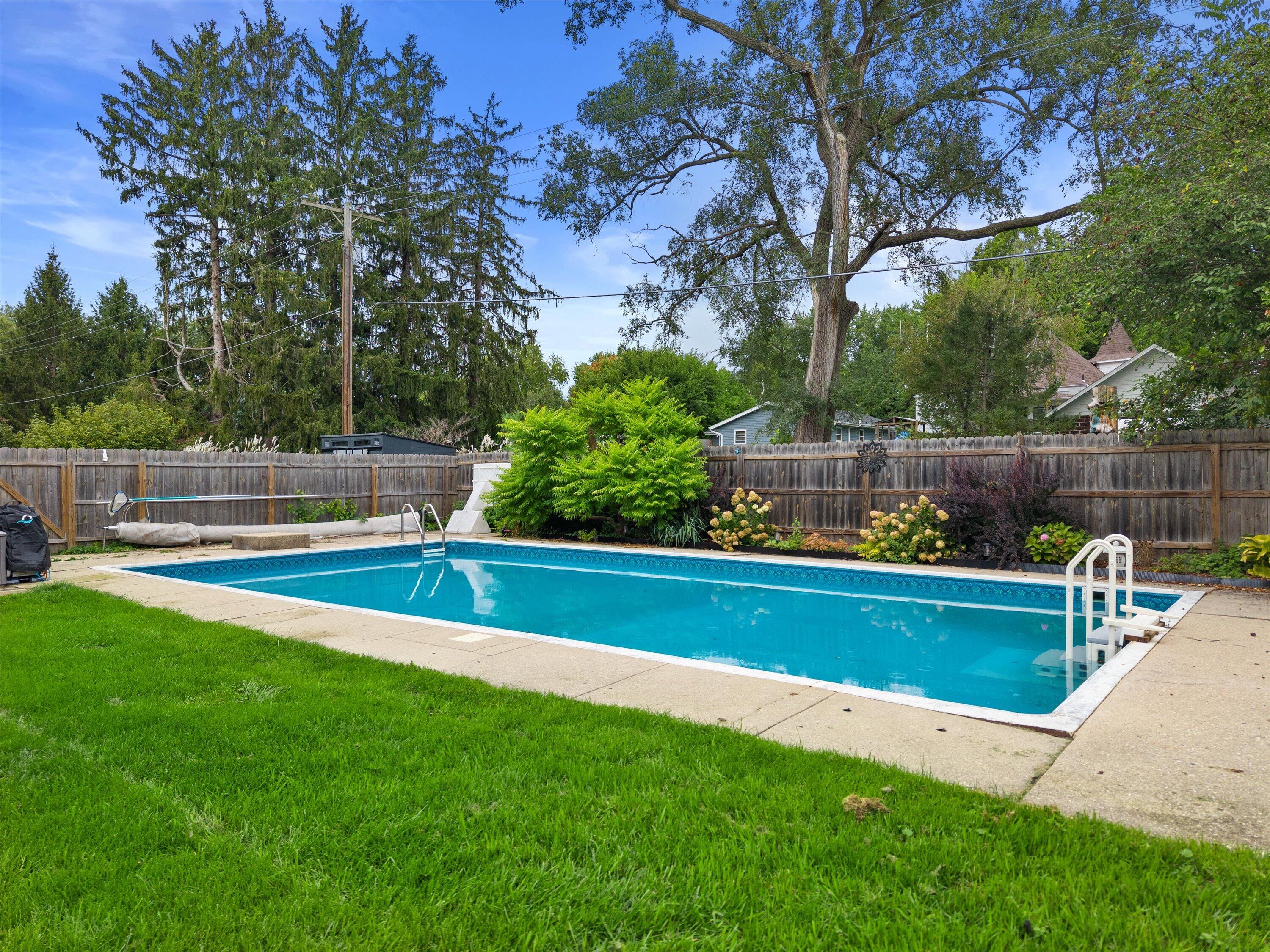 Backyard swimming pool with a concrete deck, wooden fence, and mature trees.