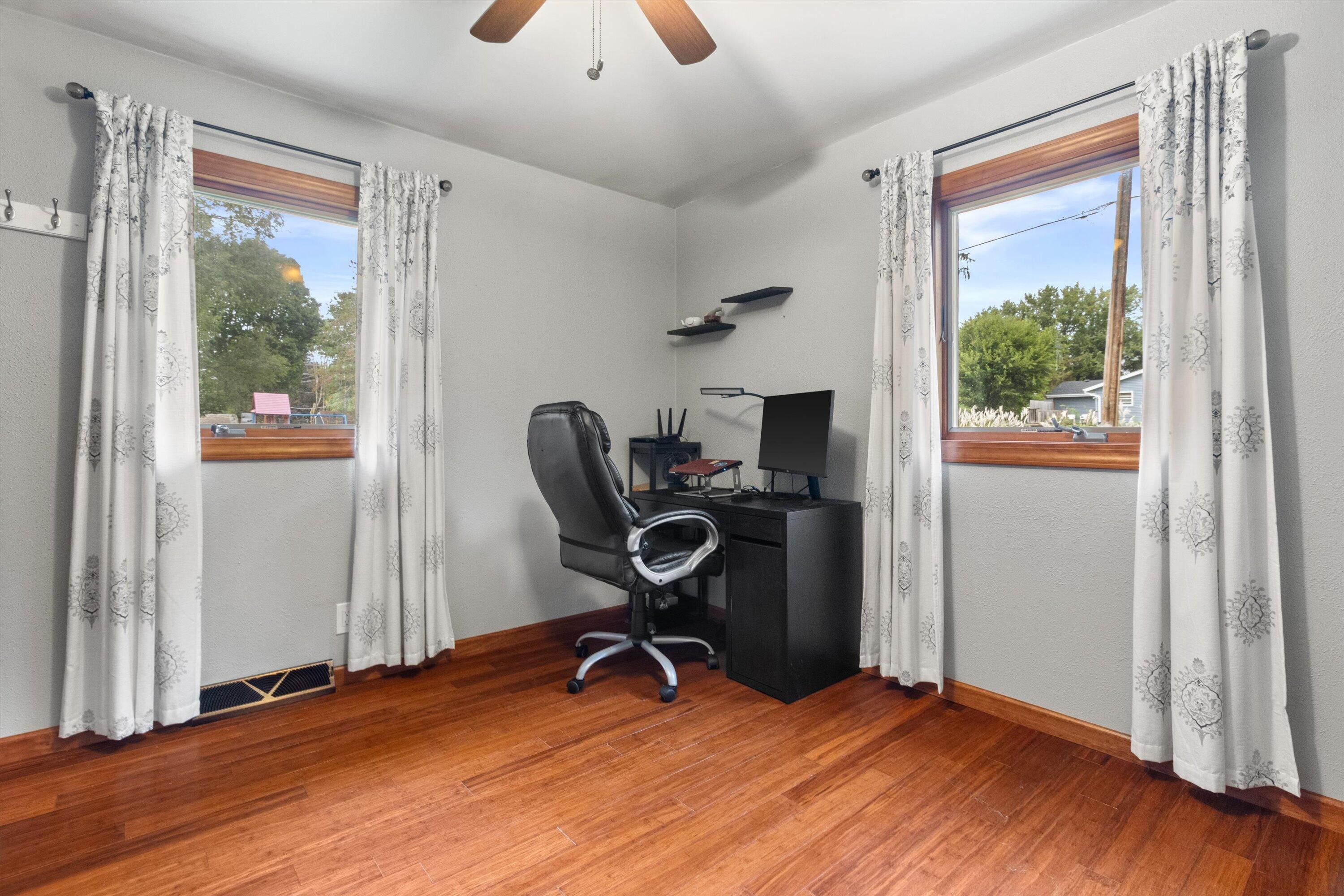 Home office with a black desk and chair between two curtained windows, above hardwood floors.