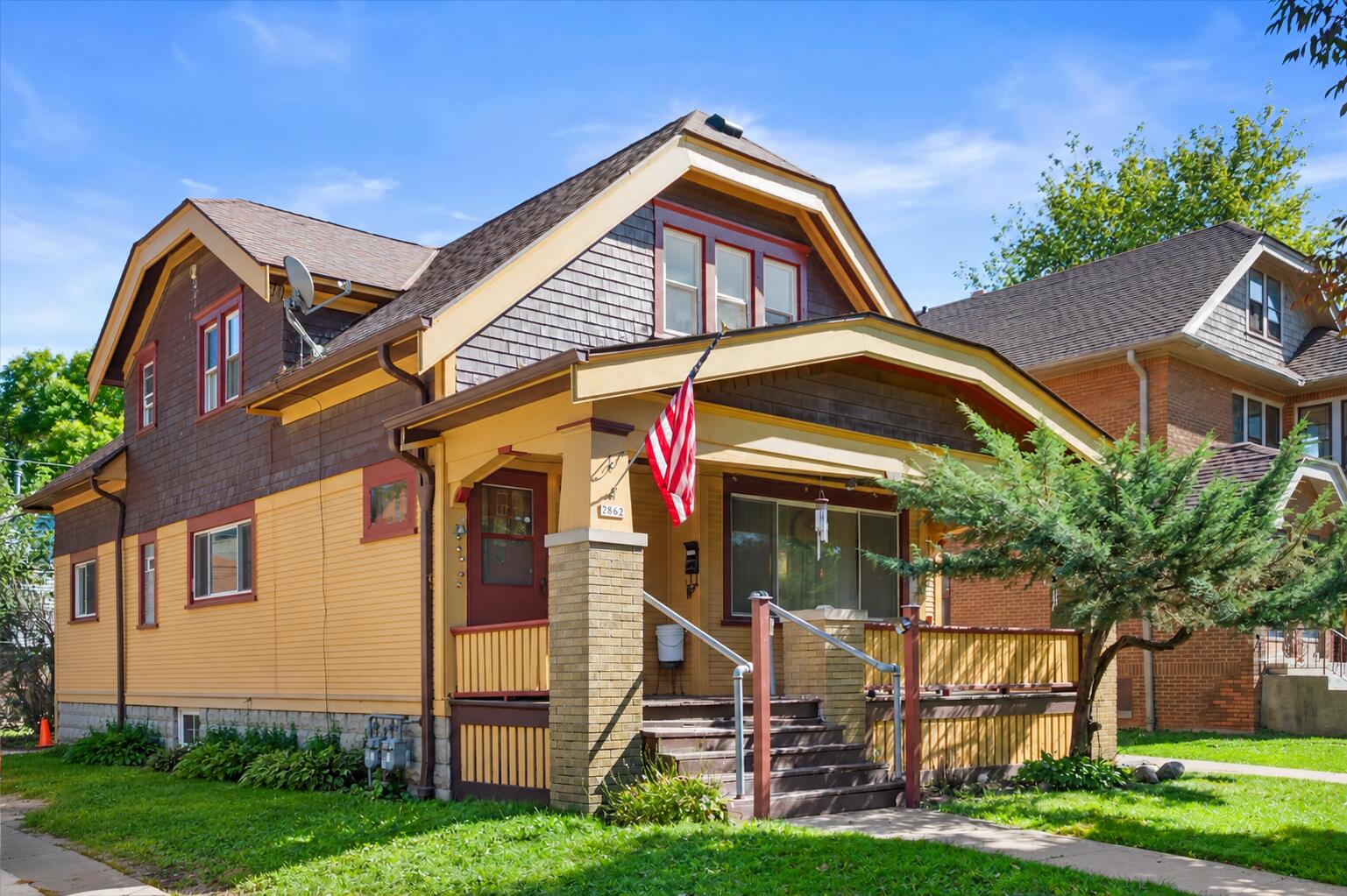 Two-story yellow house with a covered porch and an American flag beside the entrance.