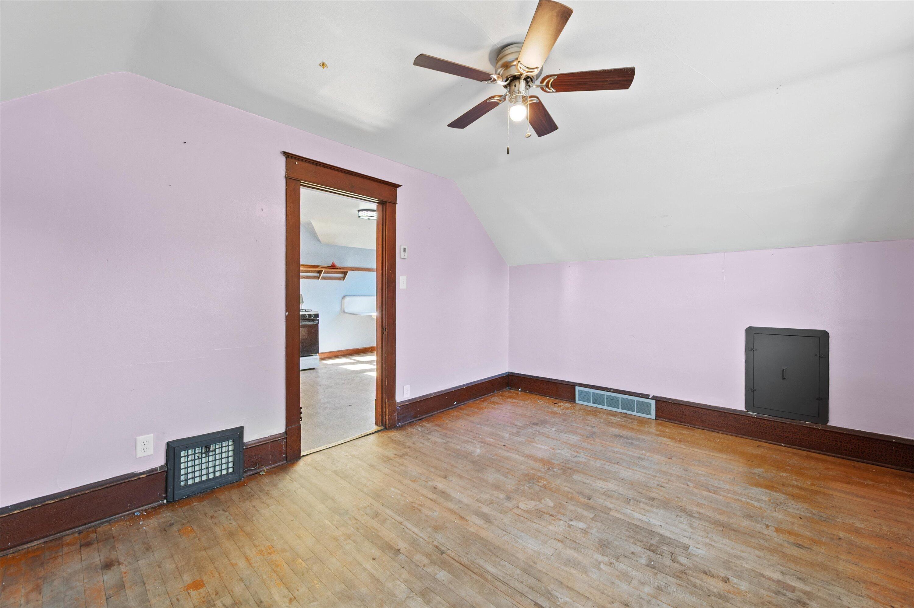 Empty lavender room with sloped ceiling, worn hardwood floors, and a ceiling fan.