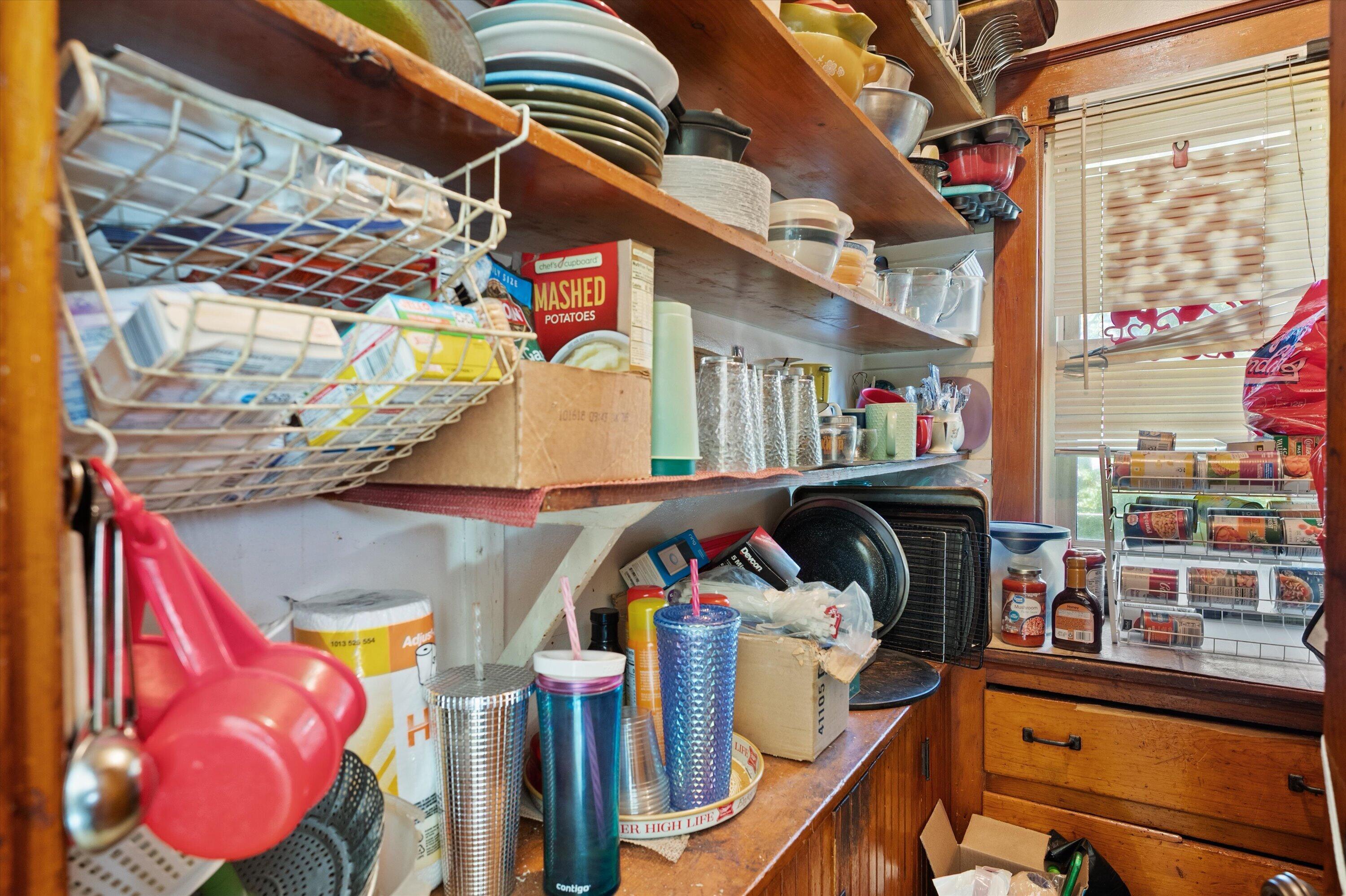 Crowded kitchen pantry with wooden shelves packed with dishes, cups, cookware, and food containers.