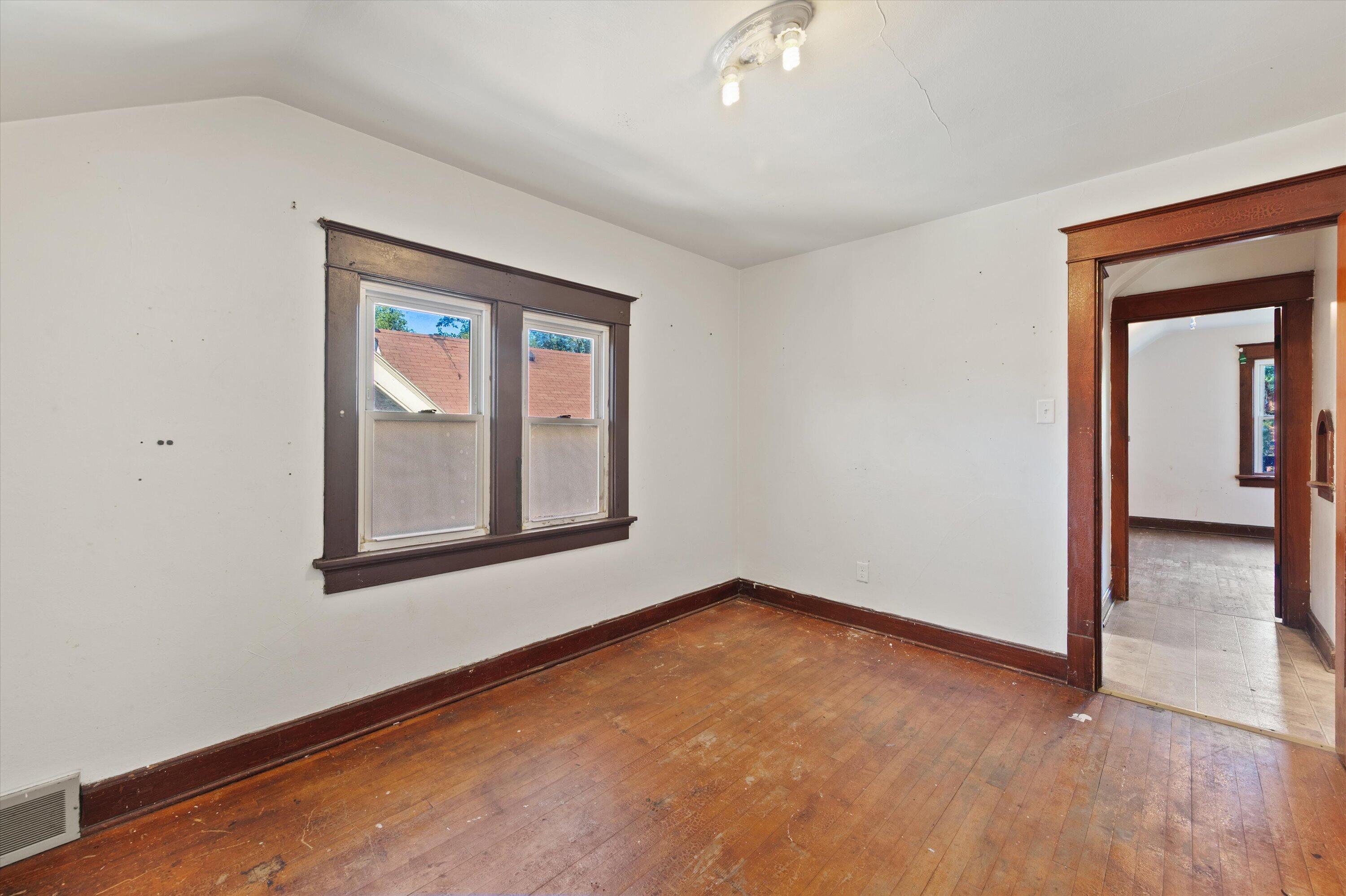 Empty room with worn hardwood floors, dark wood window trim, and a doorway to another room.