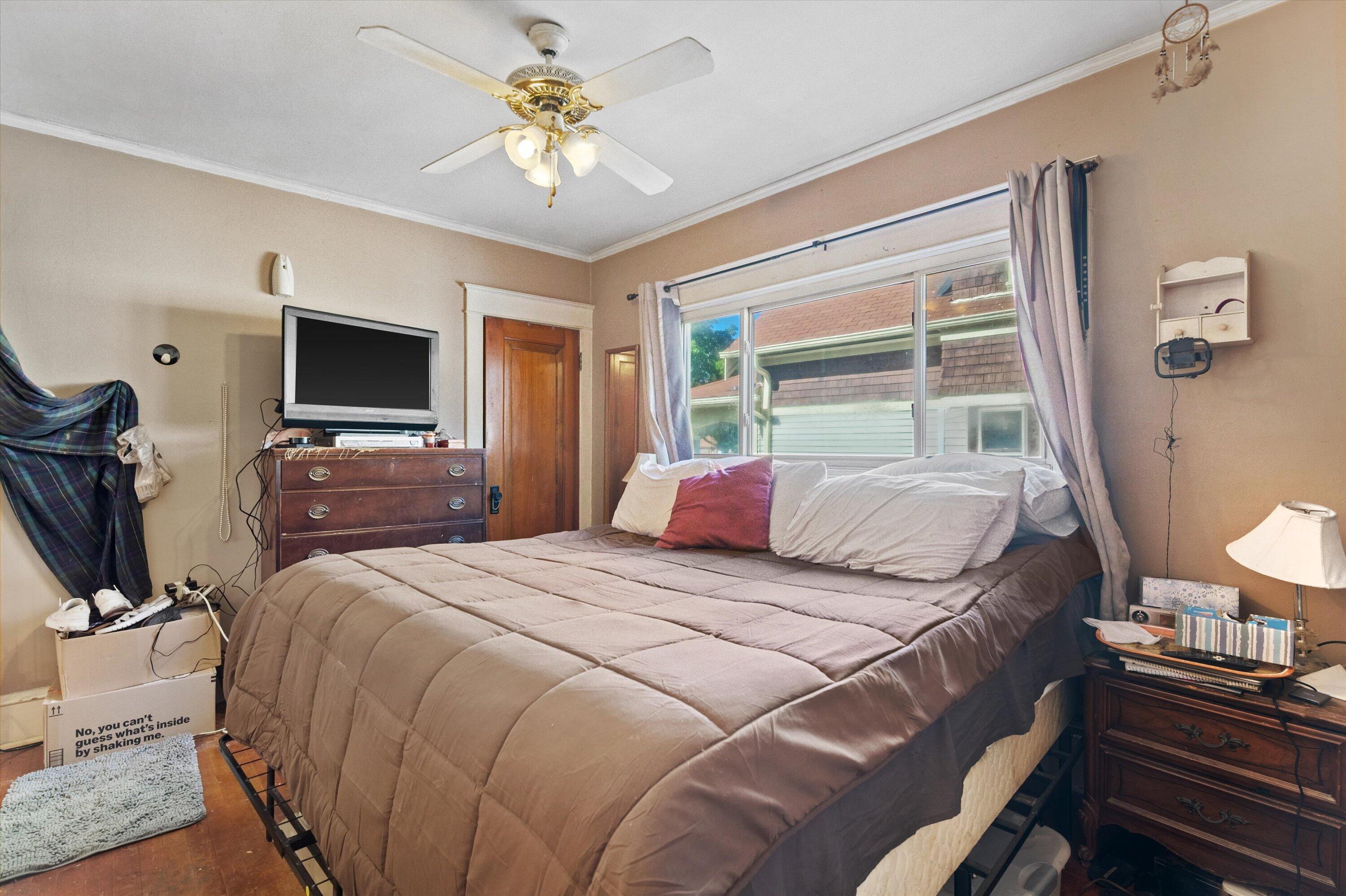 Bedroom with a large bed beneath a wide window, ceiling fan, and dresser with television.