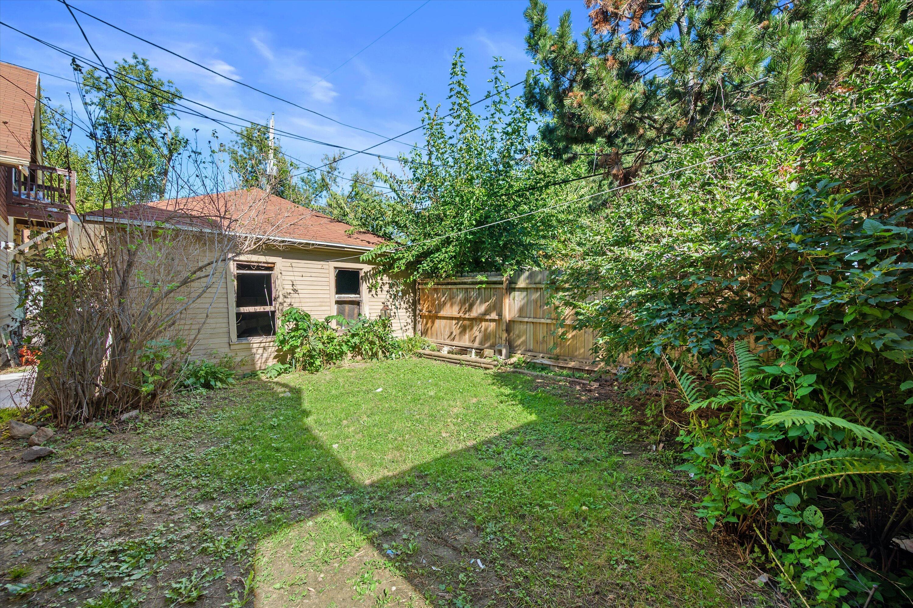 Backyard with a small lawn, dense greenery, wooden fence, and the rear of a beige house.