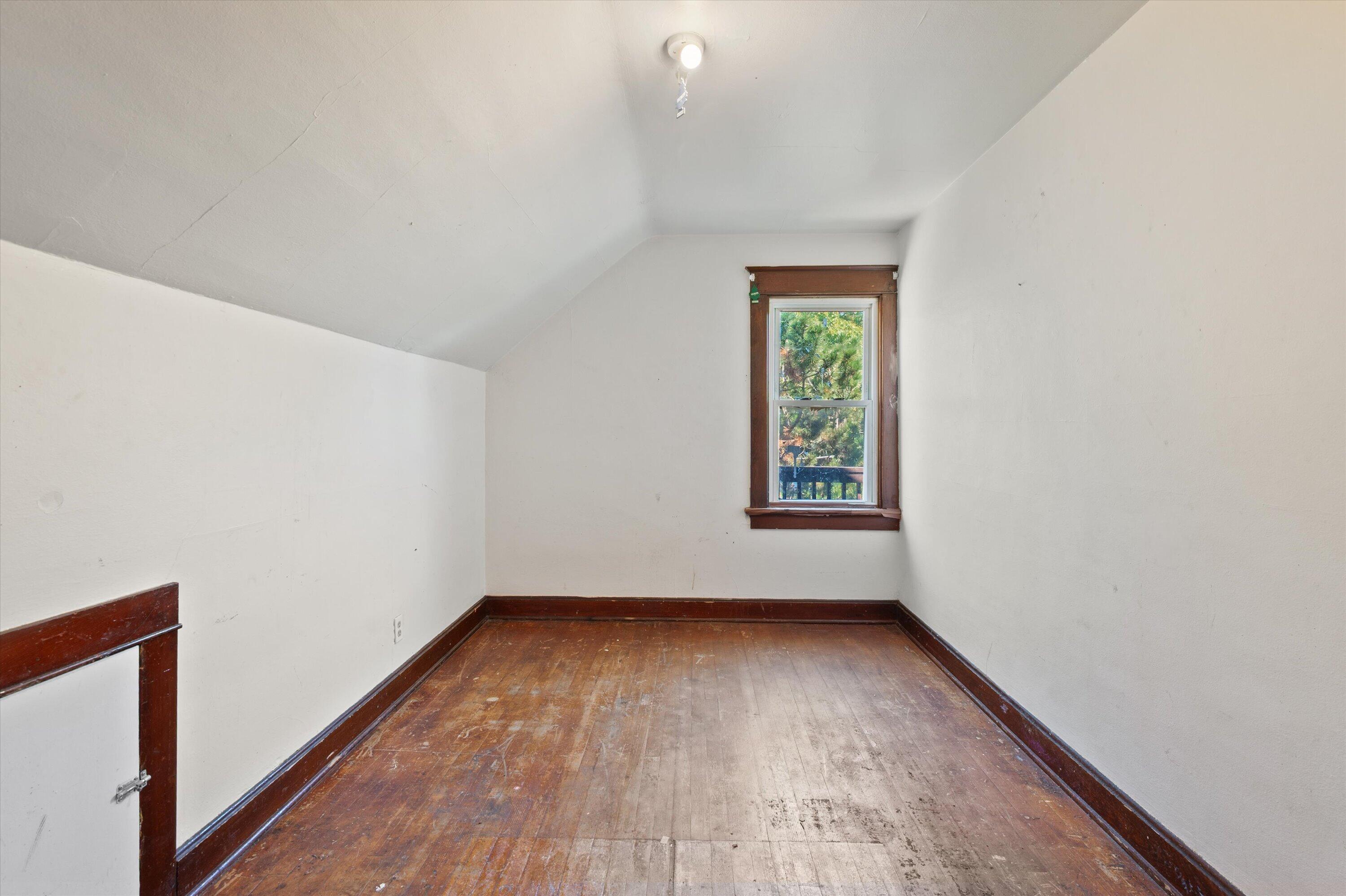 Empty room with a sloped ceiling, worn hardwood floors, and a dark-trimmed window.